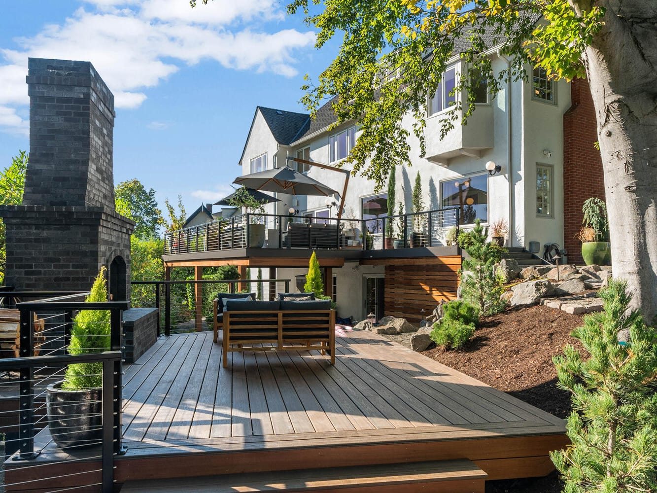 A modern multi-level deck with seating and potted plants, adjacent to a Portland, Oregon house. It features a stone fireplace and overlooks a landscaped garden. The house has large windows, and trees surround the area under a clear blue sky.