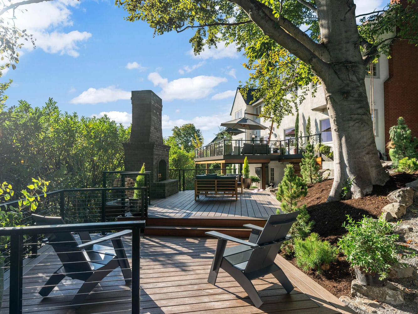 A serene backyard deck in Portland, Oregon, features wooden seating and Adirondack chairs under a large tree. The deck overlooks lush greenery and a house, with a sunny sky above. A stone chimney adds architectural interest to the landscape.