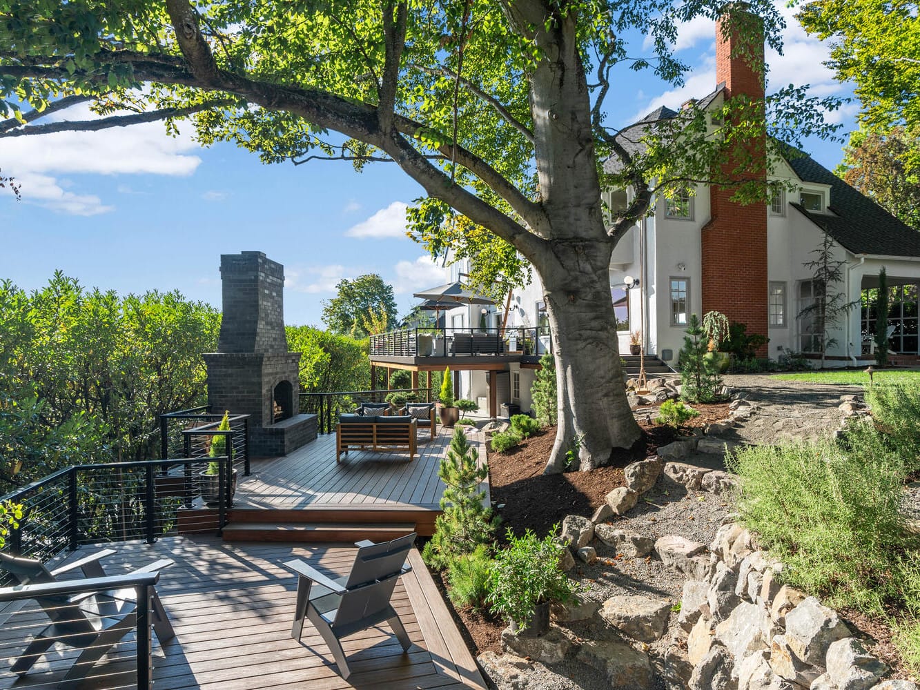 A serene outdoor scene in Portland, Oregon features a spacious wooden deck with seating under a large tree. A stone chimney stands by the deck, leading to a modern house with a red-brick chimney. The area is surrounded by lush greenery and rocks.