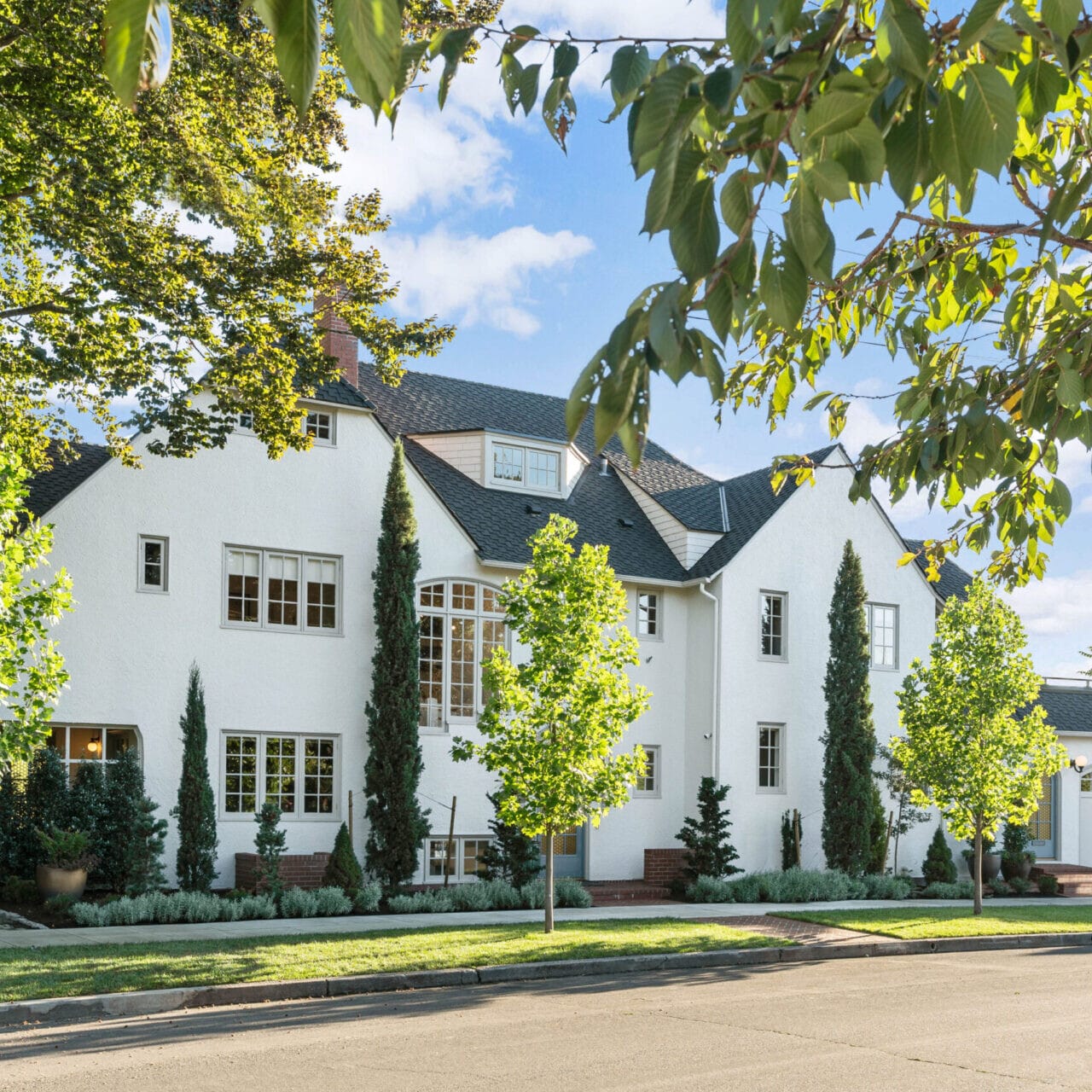 A large white house with multiple windows, surrounded by green trees and a manicured lawn. The clear blue sky enhances the serene suburban setting, showcasing Portland Oregon real estate at its finest. A driveway leads to a white double garage on the right side.