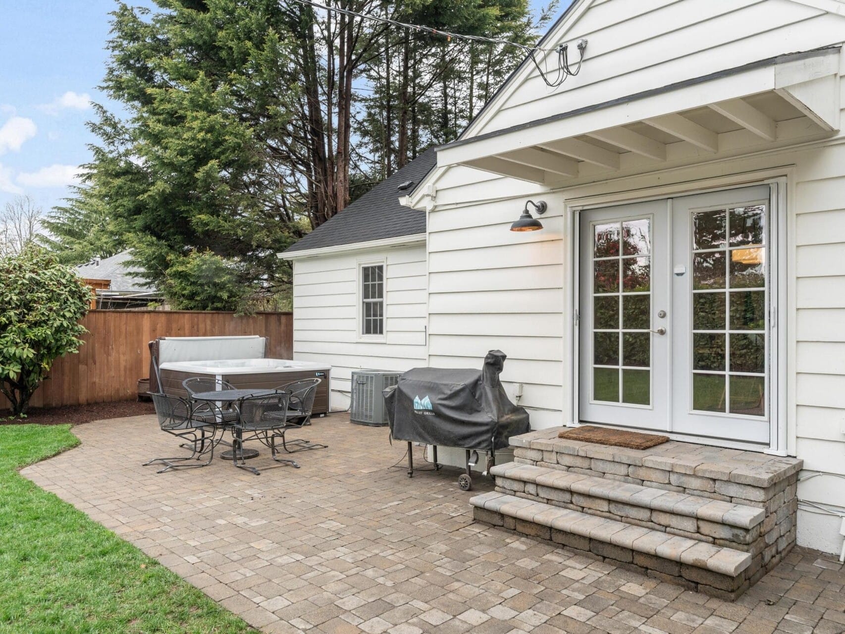 A backyard patio features a paver floor, a table with chairs, a grill under a cover, and a hot tub. The white house has French doors leading outside. A wooden fence and tall trees provide privacy. The sky is partly cloudy.