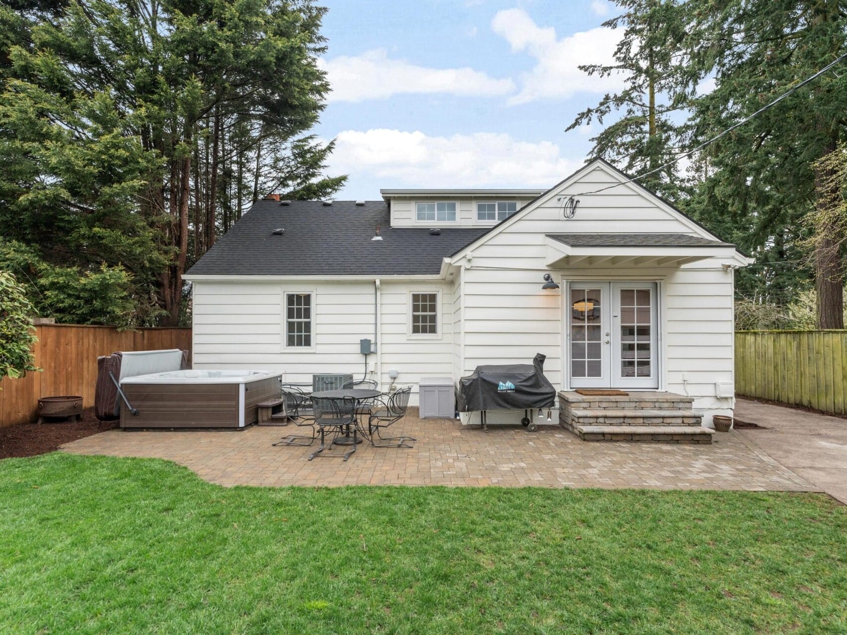 A backyard view of a white house with a paved patio, outdoor dining set, and a grill. There is a hot tub on the left. The yard is bordered by trees and a wooden fence, and the sky is partly cloudy.