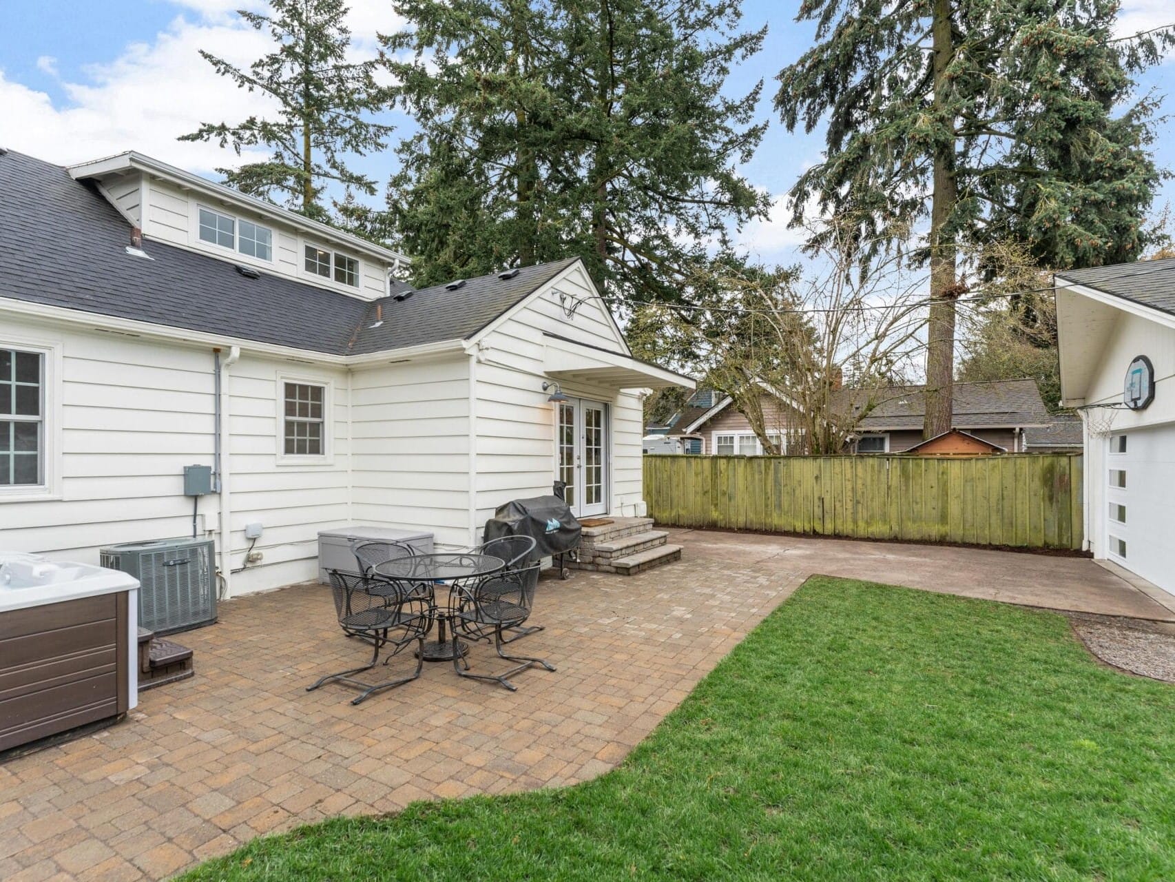 A backyard with a white house and a patio. Theres outdoor furniture, including a black metal table and chairs, a barbecue grill, and a hot tub. The area is bordered by grass and a wooden fence, with tall trees in the background.