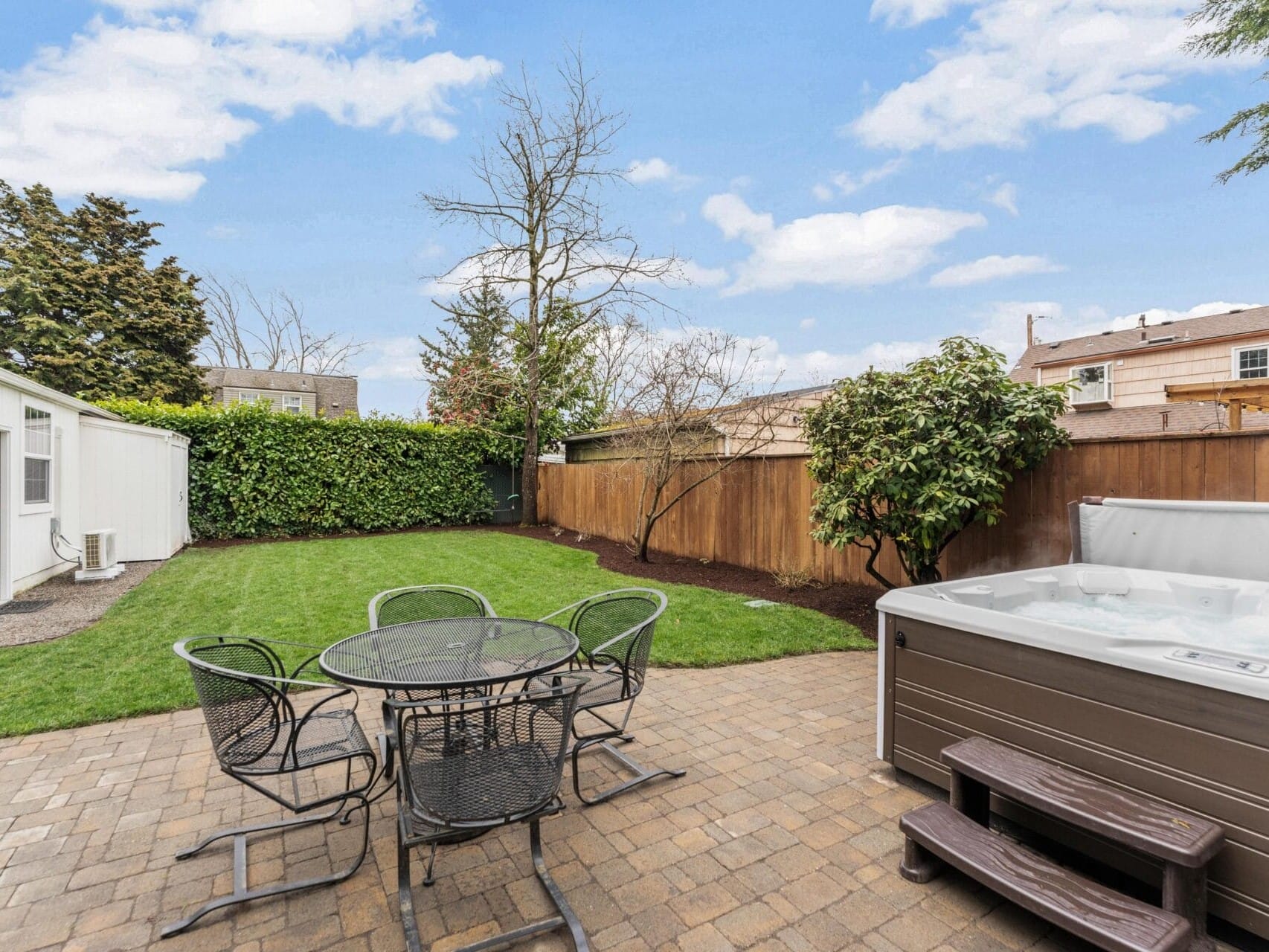 A backyard with a patio featuring a round table and chairs. A hot tub is placed nearby. Theres a green lawn, some bushes, and trees, all surrounded by a wooden fence. A shed is in the corner. The sky is clear with scattered clouds.