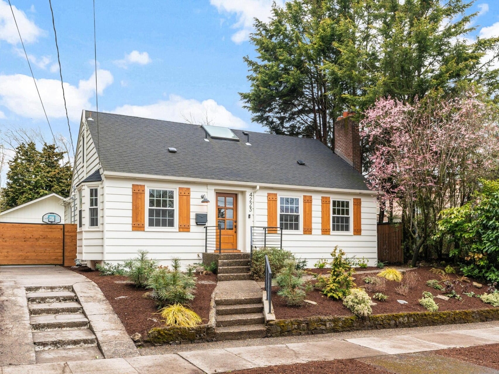 A charming white house with a dark roof, wooden shutters, and a small front porch. It features landscaped plants, a brick chimney, and a path leading to the entrance. A garage is visible in the background, surrounded by tall trees and a clear sky.