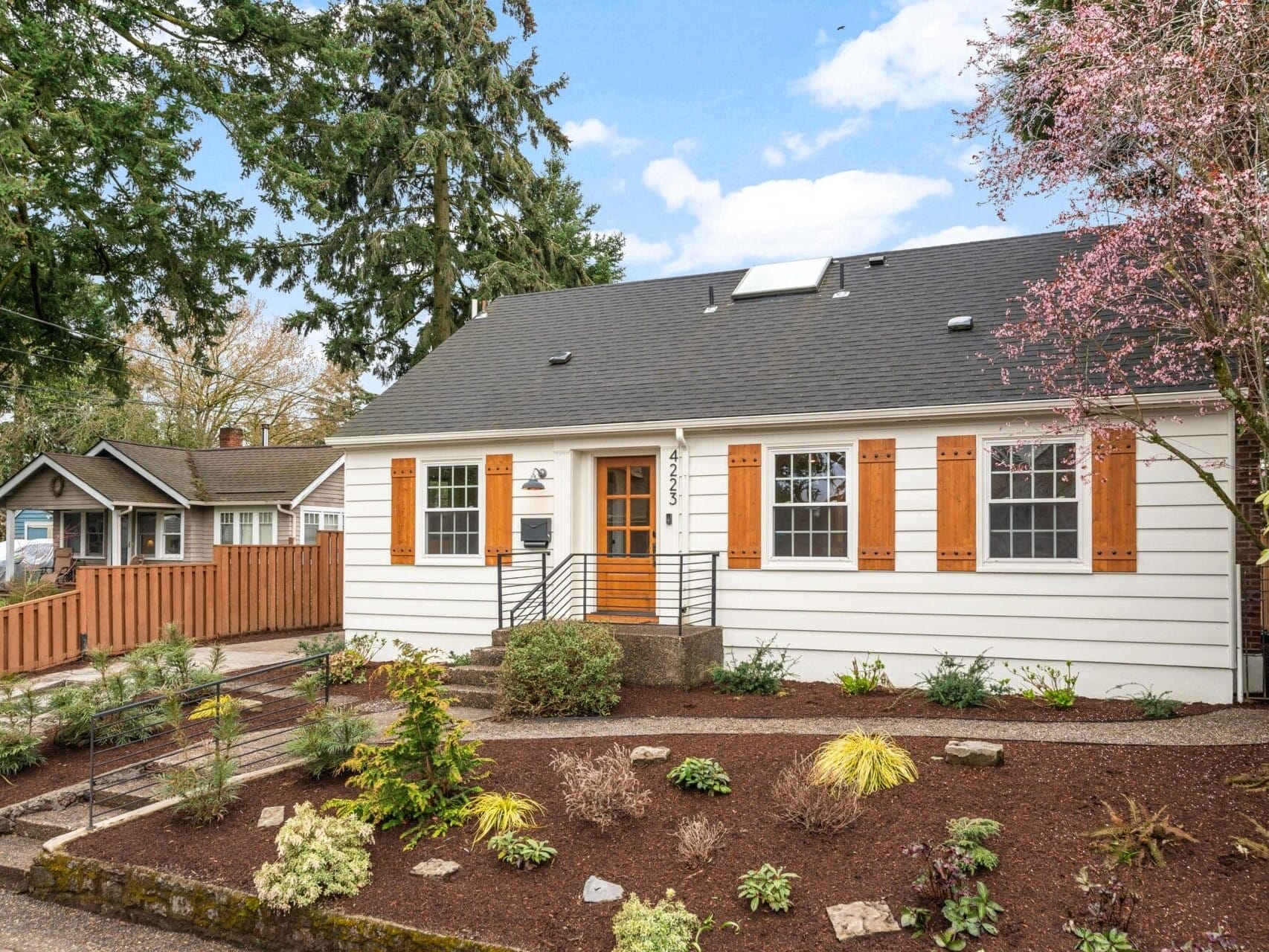 A charming white house with a black roof and wooden shutters. The front yard features a well-maintained garden with various shrubs and plants. A wooden fence lines the property, and a pathway leads to the entrance. Trees are visible in the background.