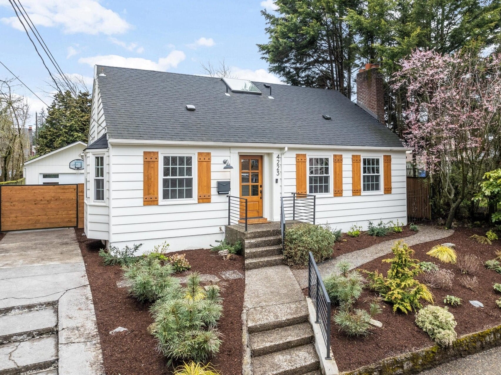 A charming white house with a dark roof, featuring orange shutters on the front windows. It has a small staircase leading to the entrance, surrounded by a manicured garden with various plants and shrubs.