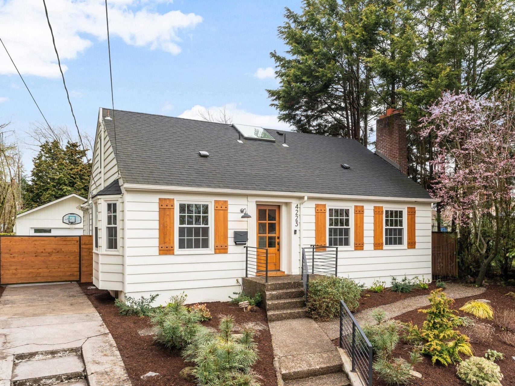 A charming white house with a gray roof, featuring wooden shutters and a central chimney. The front yard has landscaped shrubs and a concrete path leading to the entrance. Surrounded by trees under a partly cloudy sky.