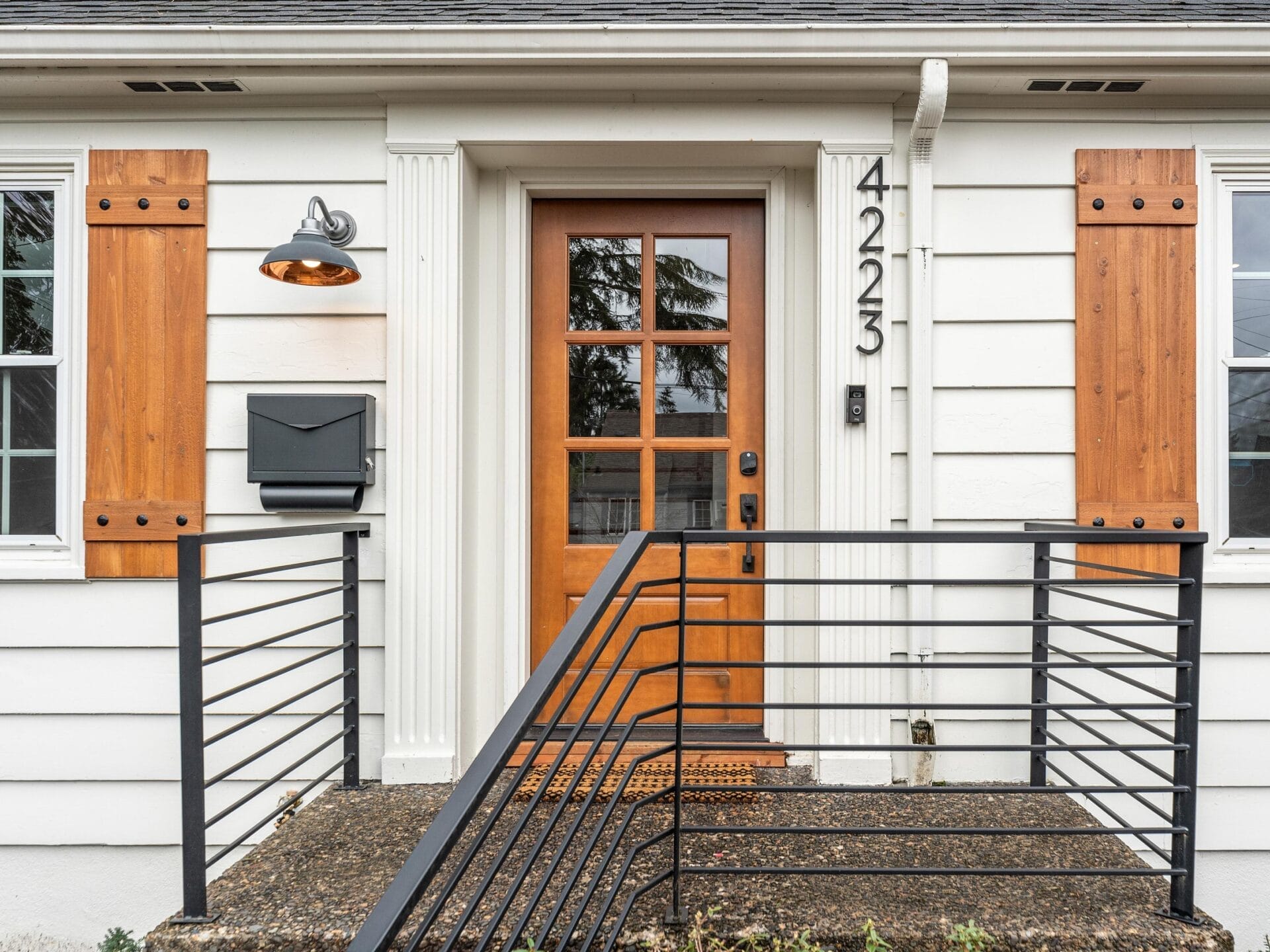 Front view of a house entrance featuring a wooden door with glass panels, framed by white siding and wood shutters. A porch light and mailbox are on the left, with black metal railing leading up stone steps. House number 4223 is displayed vertically.