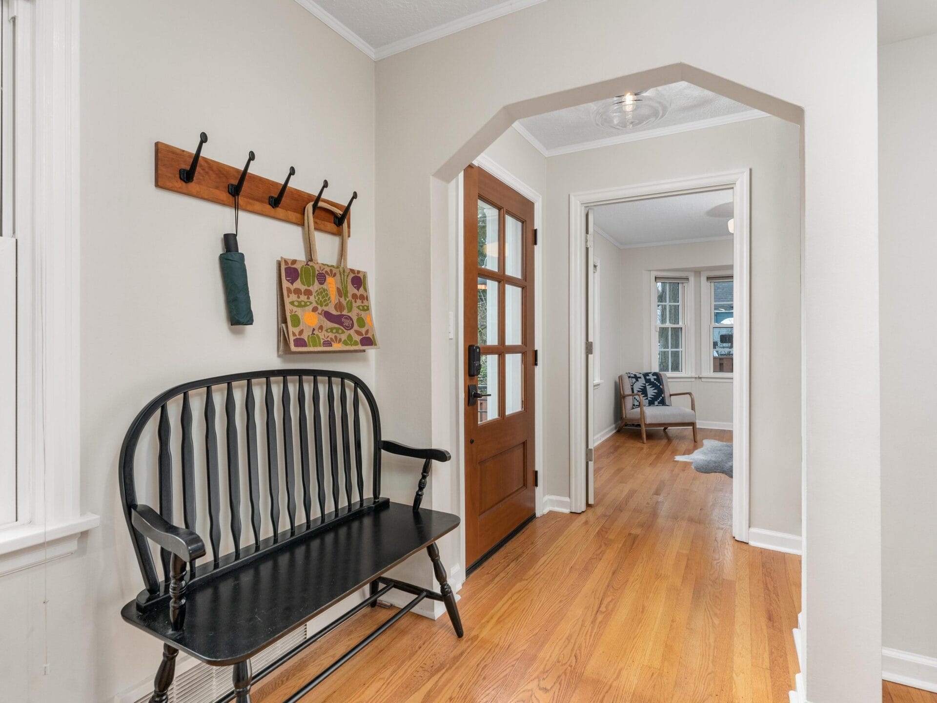 A cozy entryway features a wooden floor and arched doorway leading to a bright room. A black bench with a wooden coat rack above holds bags and an umbrella. A wooden door with glass panels adds warmth to the space. A chair is visible in the adjacent room.