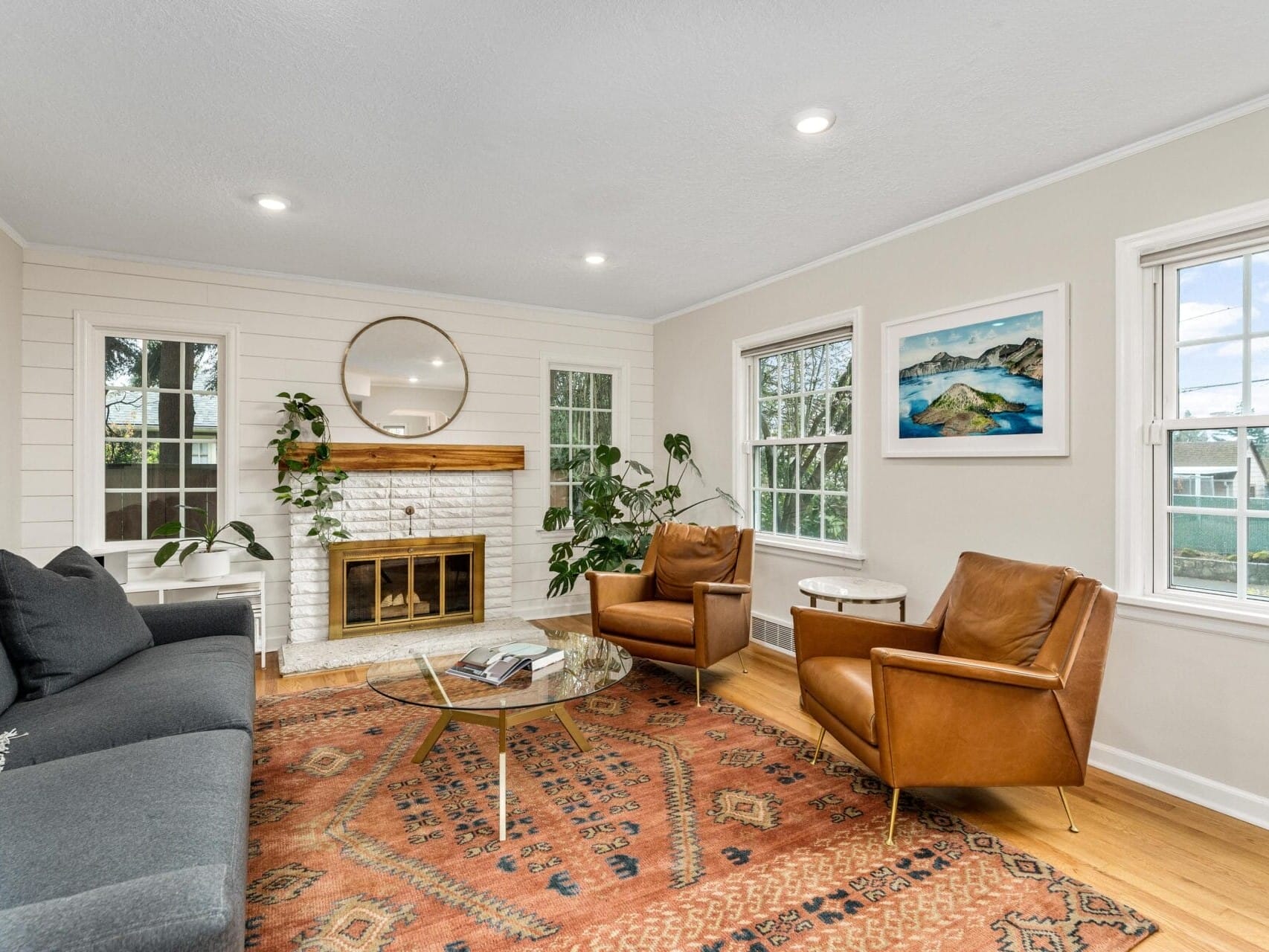 A cozy living room with a white brick fireplace, two brown leather chairs, a gray sofa with yellow pillow, and a round glass coffee table on a patterned rug. Large windows illuminate the space, and a round mirror hangs above the mantel with plants.