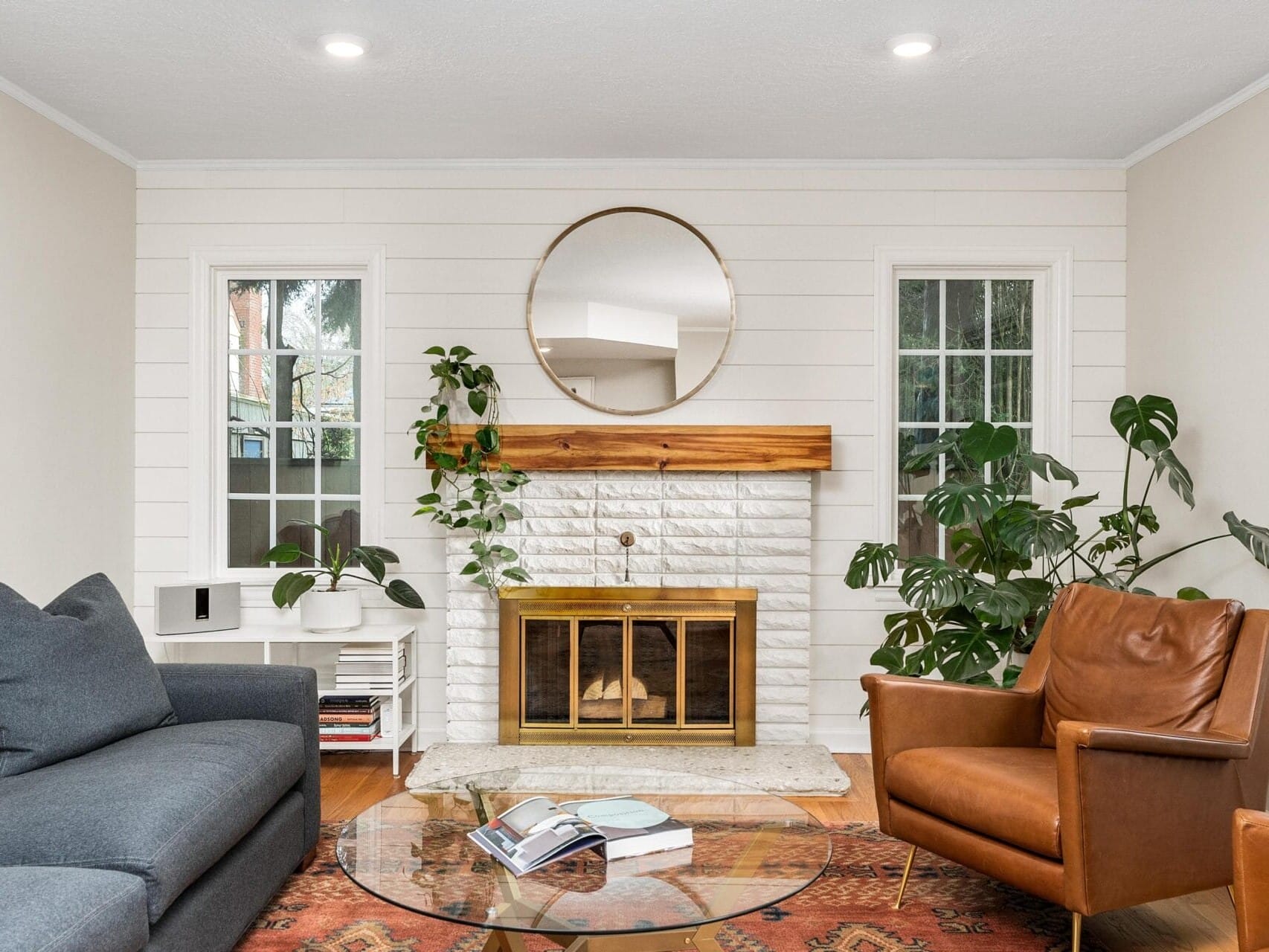 A cozy living room with a white brick fireplace, round mirror above, and potted plants on each side. A gray couch and a brown leather armchair surround a glass coffee table atop a patterned rug. Natural light streams in through the windows.