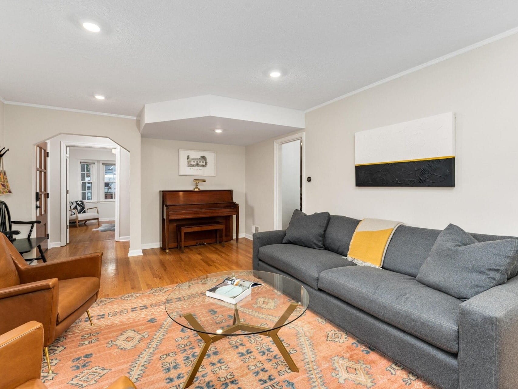 A cozy living room with a gray couch and yellow throw blanket, two brown leather chairs, a glass coffee table on a patterned rug, a piano by the wall, and a small round table with a chair in the corner. Walls are light-colored with a neutral painting.