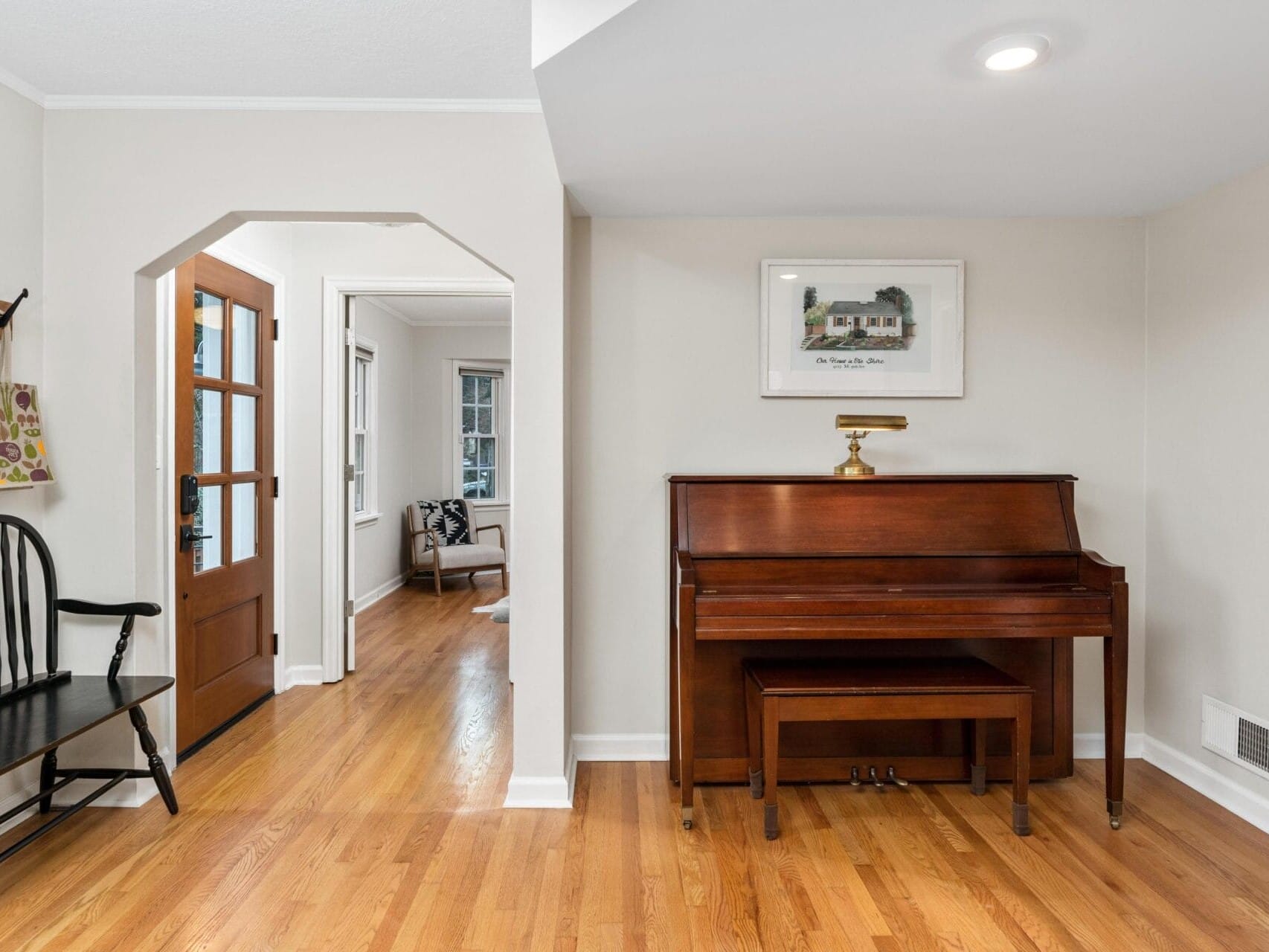 A cozy interior features a wooden piano with a bench against the wall, beneath a framed painting. To the left, theres a wooden chair and a coat rack with a bag. A doorway opens to another room with a chair. Light hardwood flooring throughout.