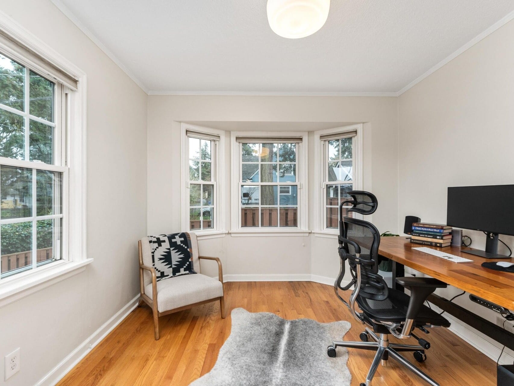 A home office with a wooden desk, ergonomic chair, computer monitor, and books. A patterned chair sits nearby. Windows with a view of a fenced backyard and trees let in natural light. A light fixture hangs from the ceiling.