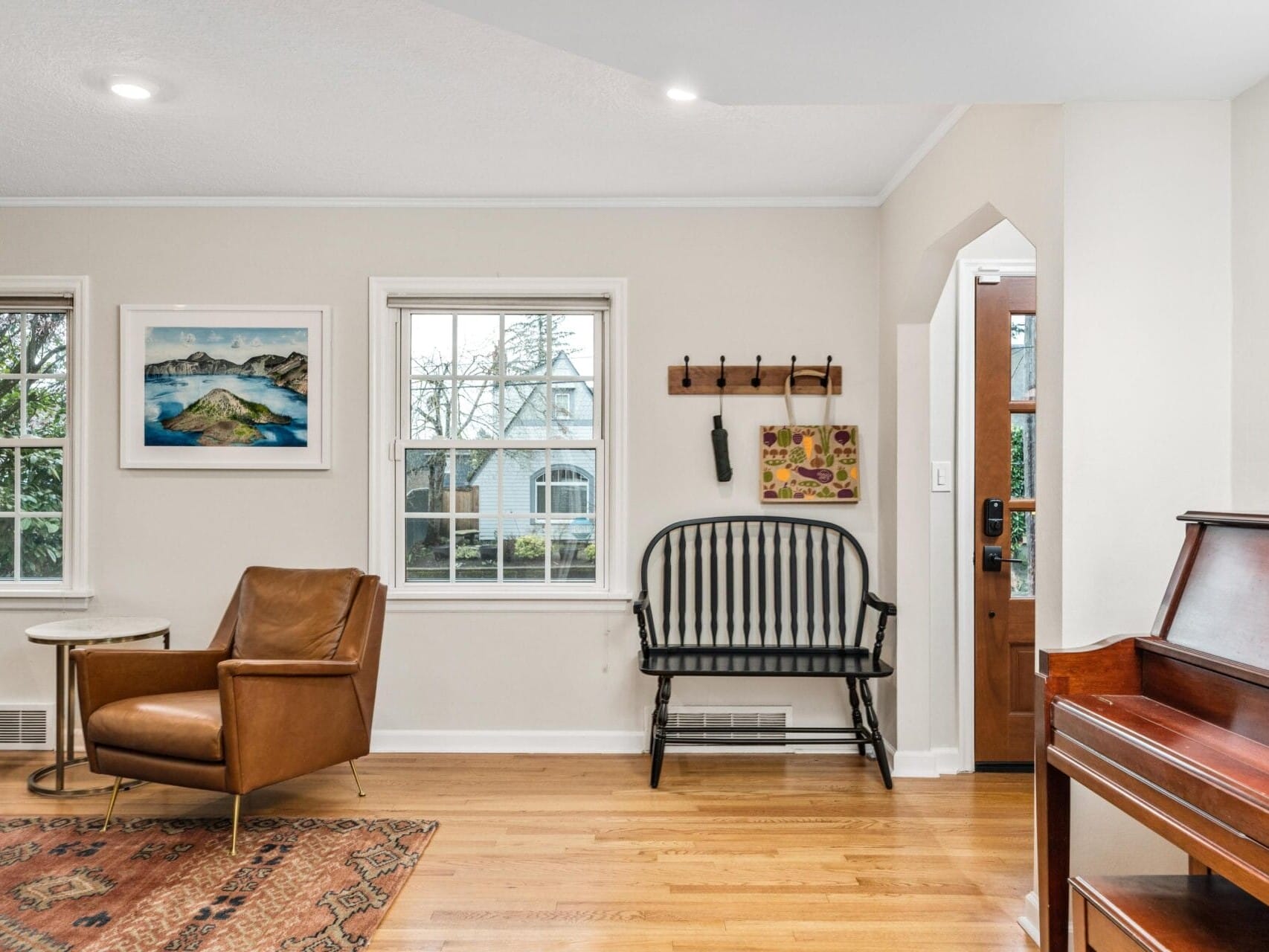 A cozy living room with a leather armchair, a wooden bench, and a piano. Two windows display a scenic view. A framed picture hangs on the wall, and a small side table with a rug completes the setting, alongside a coat rack near the entrance.