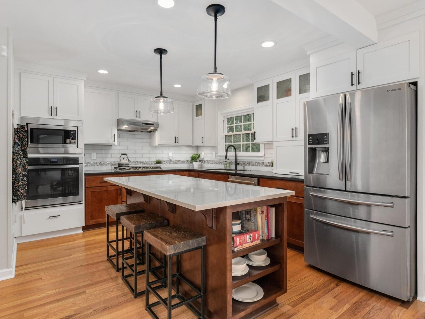 Modern kitchen with white cabinets, stainless steel appliances, and wooden flooring. A large island with a gray countertop features two wooden stools and built-in shelves. Pendant lights hang overhead, and a window allows natural light in.