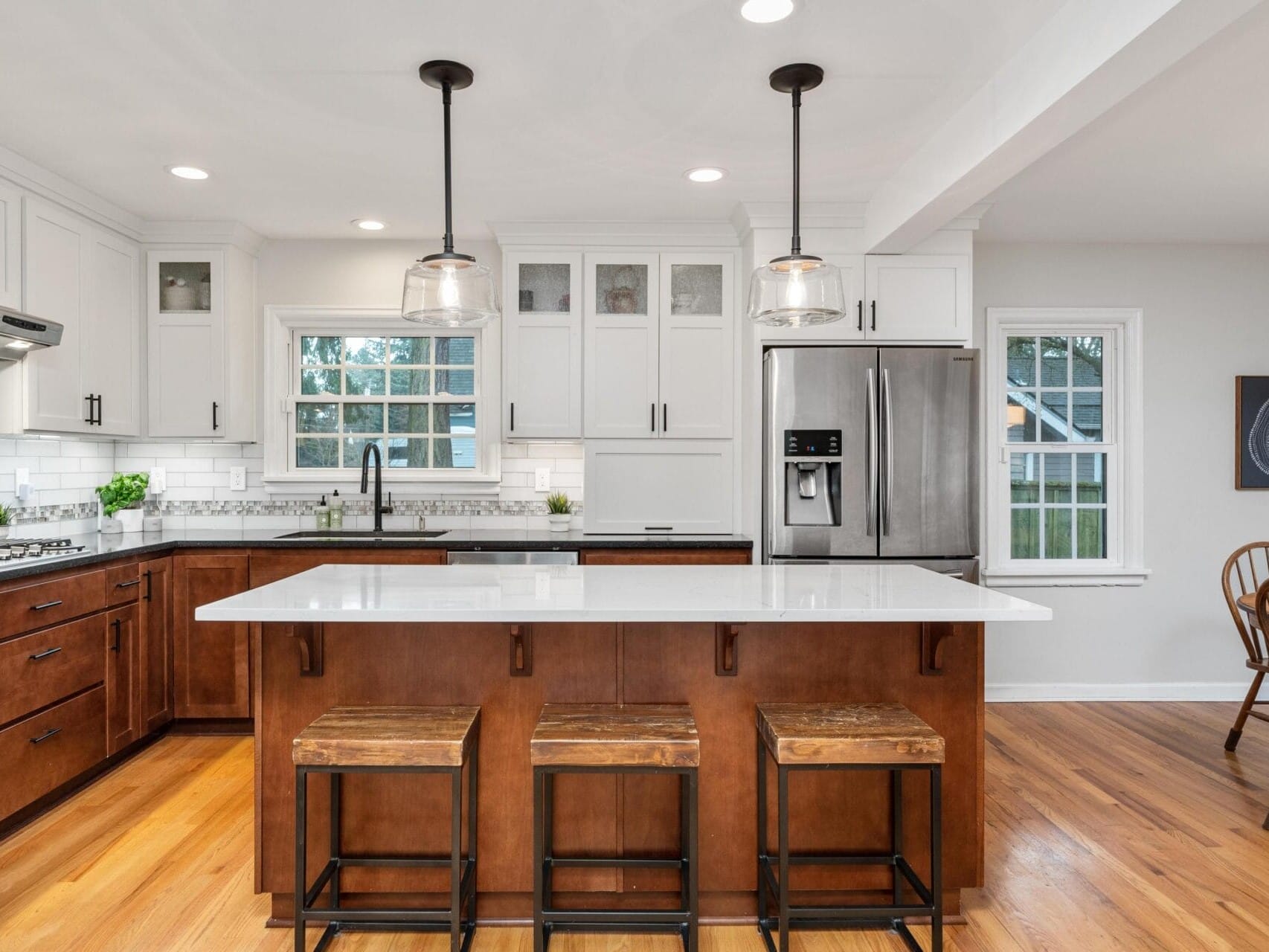 Modern kitchen with wooden floors, a large white island with three wooden stools, and stainless steel appliances. Hanging pendant lights illuminate the space. White cabinets and a small dining area with a wooden table and chairs are visible.