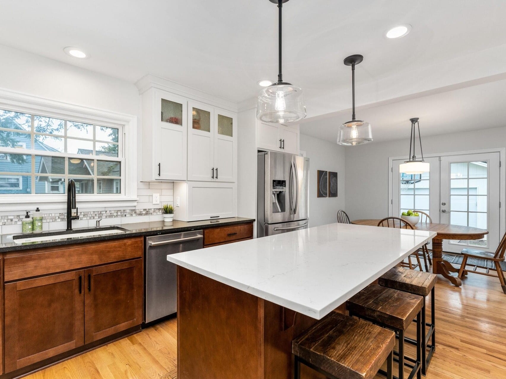 A modern kitchen with wooden cabinets, white countertops, and a central island with stools. Stainless steel appliances include a refrigerator and dishwasher. Pendant lights hang above the island, and a dining table is visible in the background.