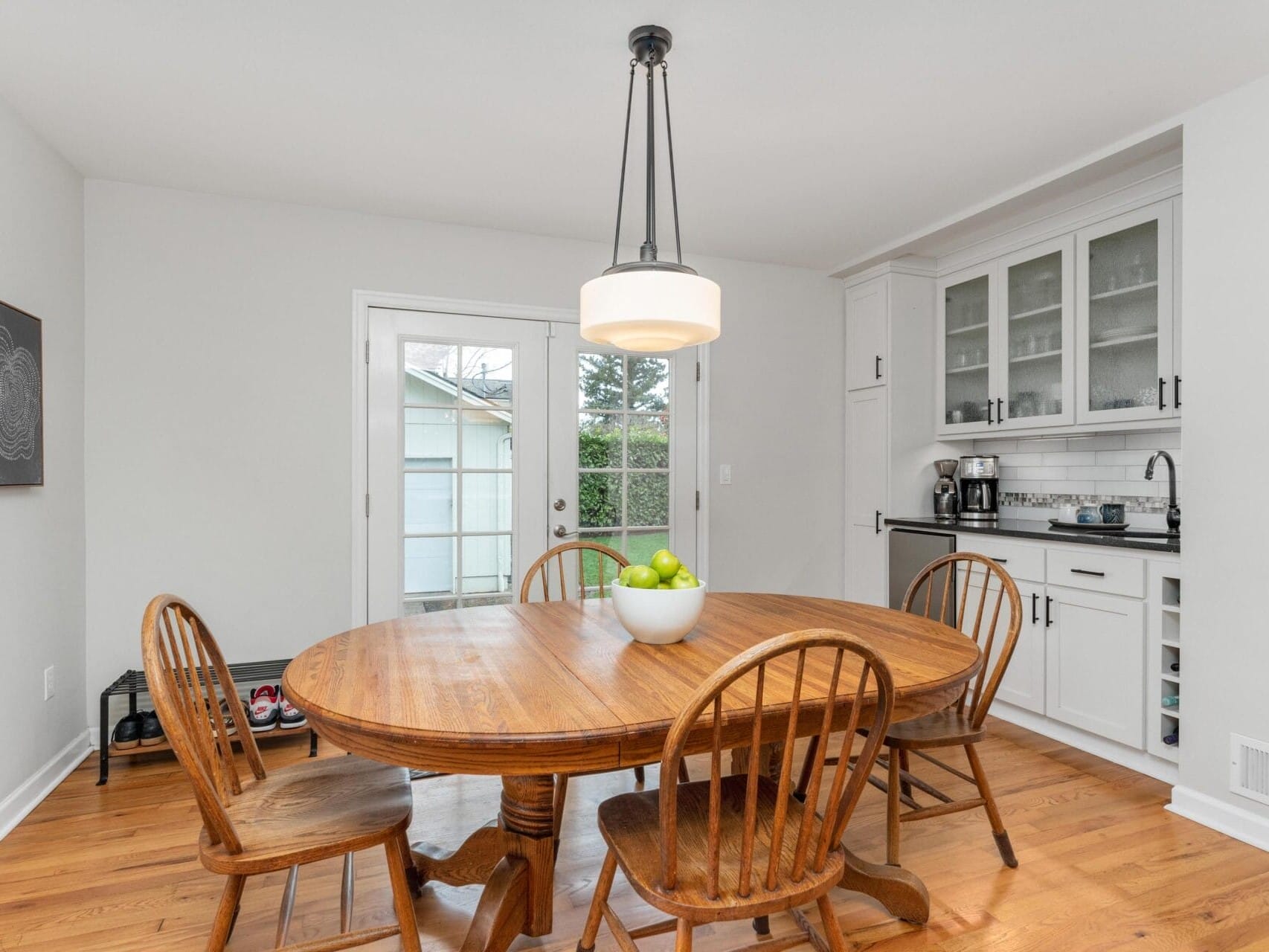 A cozy dining area with a wooden table and four chairs, a bowl of green apples centered on the table. Glass doors in the background lead outside. A white cabinet with glass doors and a small counter with a coffee maker is on the right.