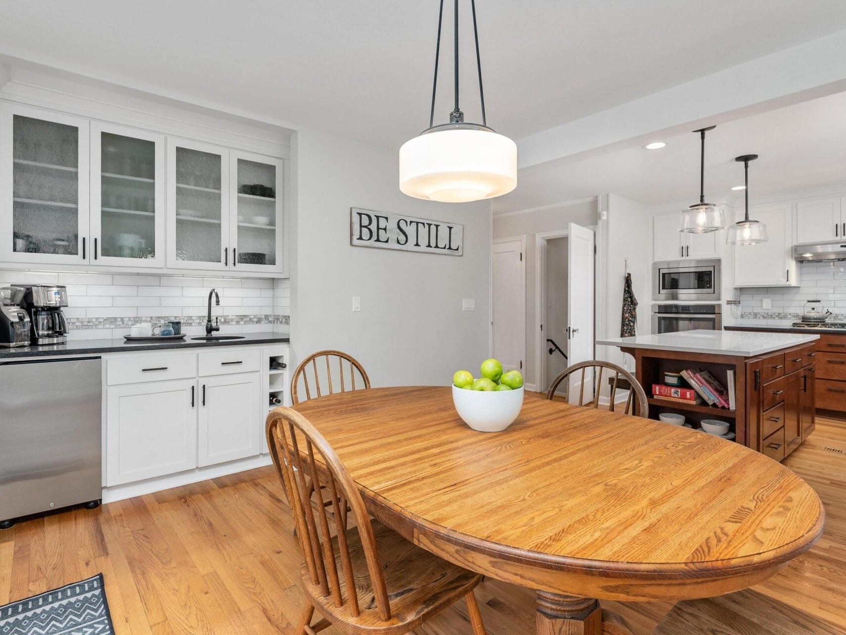 A bright kitchen with wooden floors and an oval wooden dining table with chairs. The table has a bowl of green apples. The kitchen features white cabinets, a coffee maker, sink, stove, and an island. A Be Still sign is on the wall. Pendant lights hang above.