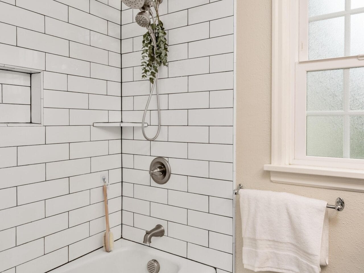 A clean bathroom features white subway tiles and black hexagonal floor tiles. A white bathtub has a silver showerhead above, with some green foliage hanging. A towel hangs on a rack beneath a white-framed window.