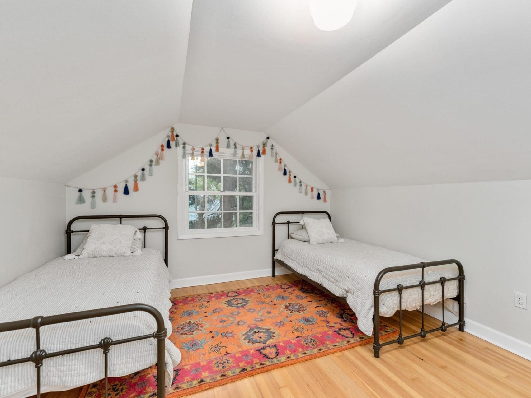 A cozy attic bedroom featuring two twin beds with metal frames, white bedding, and a colorful patterned rug. A string of decorative tassels hangs across the sloped ceiling above a window bringing in natural light.