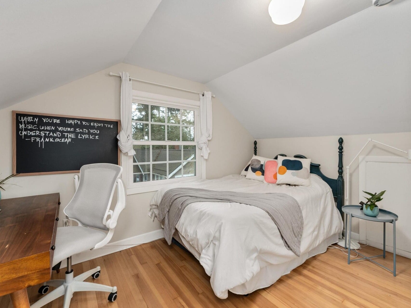 A cozy bedroom with a bed under a window, adorned with a gray blanket and decorative pillows. A chalkboard on the wall has writing on it. A wooden desk with a white chair is in the left corner. Two small plants add a touch of greenery.