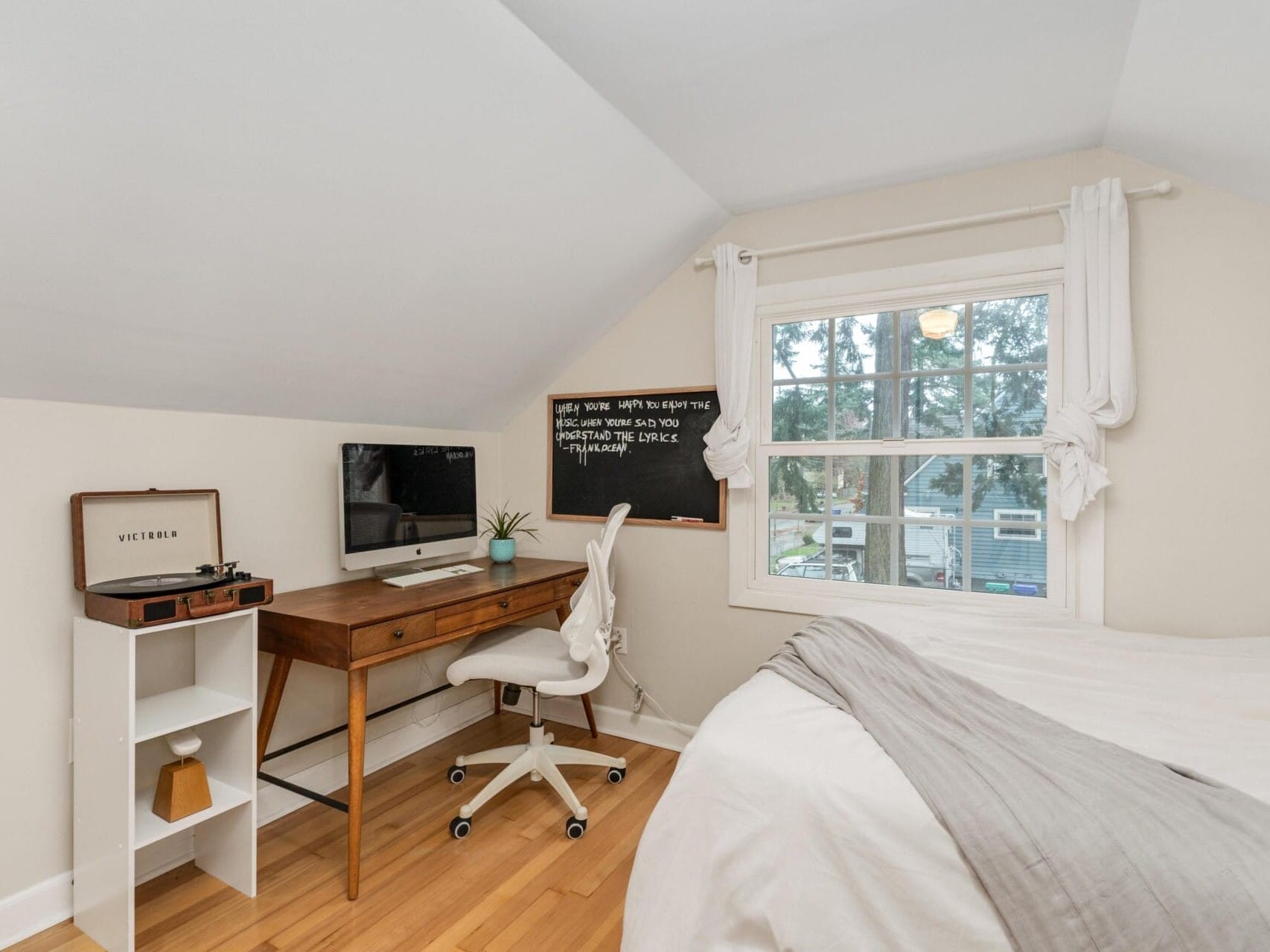 A cozy bedroom with a sloped ceiling features a wooden desk with a computer and a chalkboard. A turntable sits on a small shelf. A bed with a gray blanket is next to a window overlooking green trees.