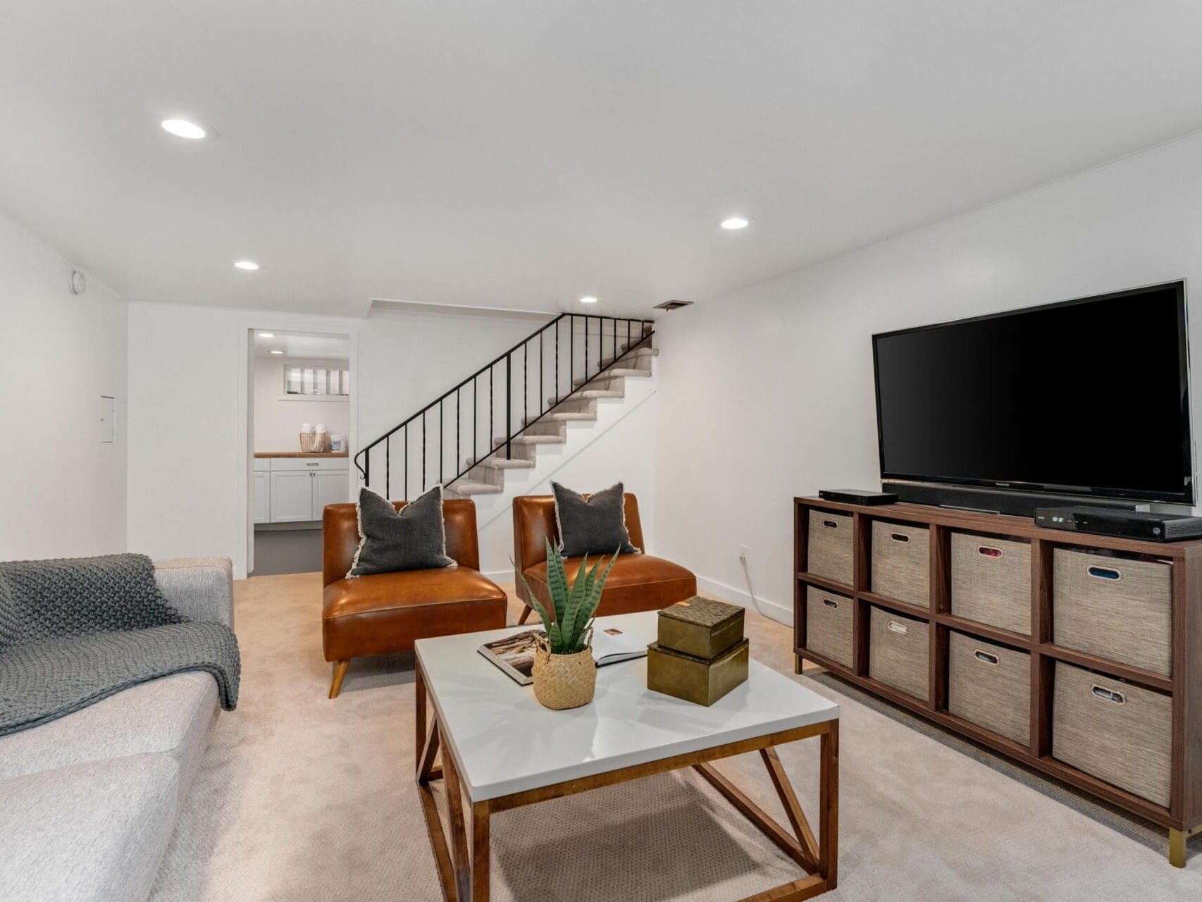 A cozy living room with a white sofa, two brown leather chairs, and a wooden coffee table with a plant. A large TV sits on a unit with storage baskets. The room leads to a staircase and a small kitchenette in the background. Bright, modern decor.