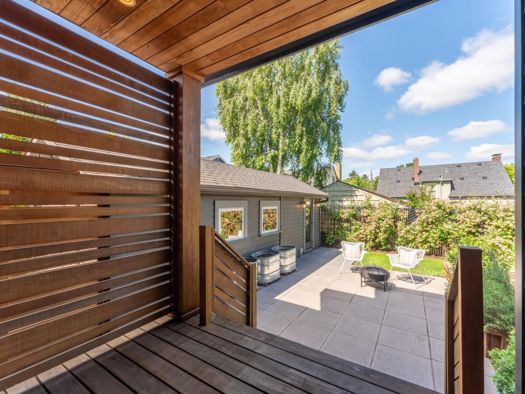 A modern terrace with wooden paneling overlooks a sunny patio featuring two white chairs and a small table. In the background, a single-story house sits adjacent to a hedge lined with greenery, and the sky is bright and partly cloudy.