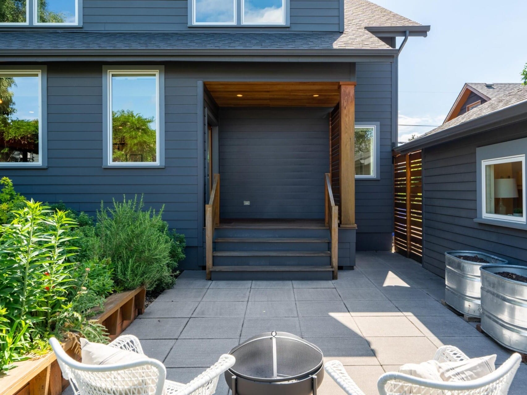 A modern gray house exterior with a small porch and wooden steps. The patio area features white wicker chairs and a round fire pit, surrounded by greenery and plants in wooden planters. Bright, sunny day with a clear blue sky.