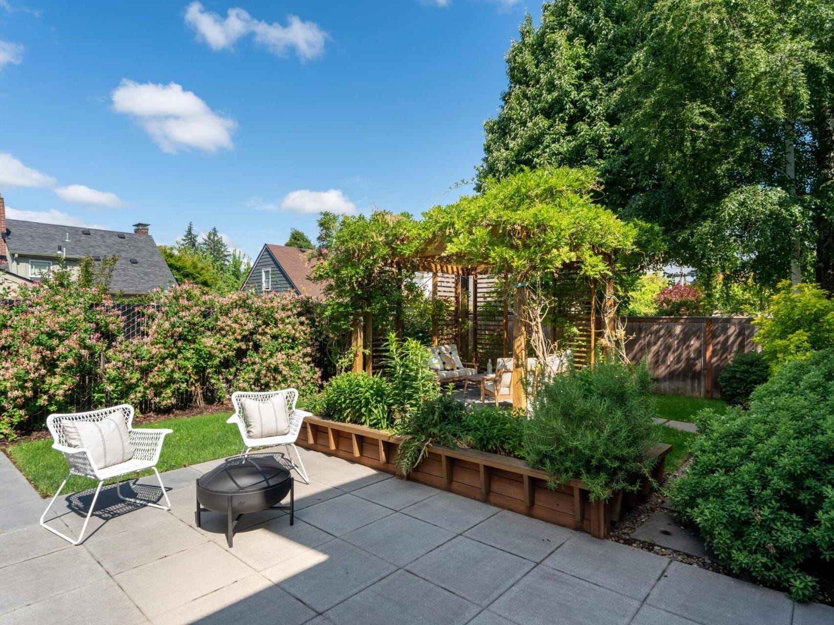 A sunny backyard features a paved patio with two white chairs and a small black fire pit. A lush garden surrounds the area, and a wooden pergola with greenery offers shade. Tall trees and a wooden fence form the backdrop under a clear blue sky.