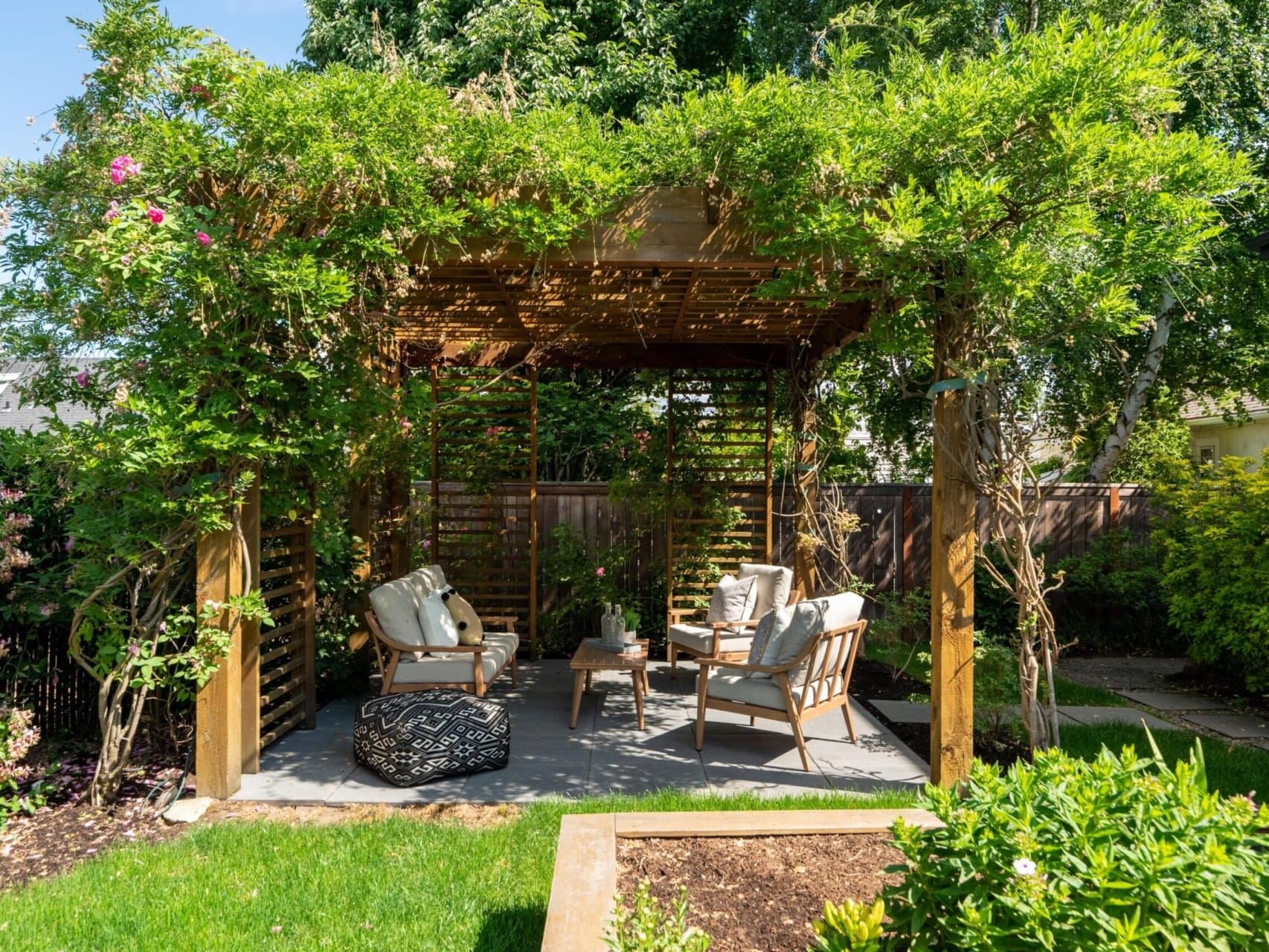 A wooden pergola adorned with lush greenery and vines shelters cushioned seating and a small table in a garden. Bright sunlight filters through the leaves, casting shadows on the grass and creating a serene outdoor space.