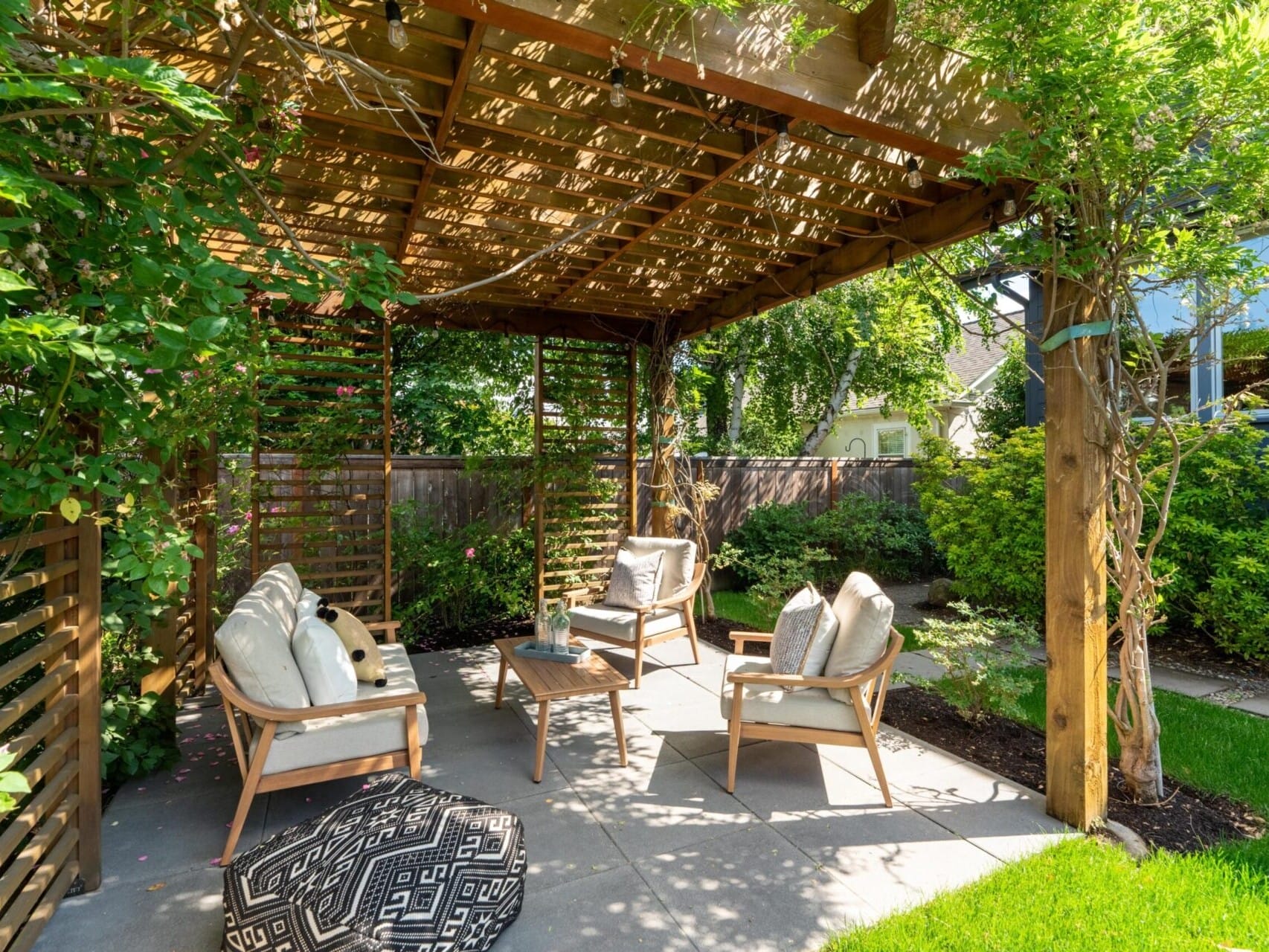 A shaded wooden pergola in a garden, with four cushioned chairs surrounding a small table on a stone patio. The pergola is adorned with climbing plants, and the area is surrounded by lush greenery.