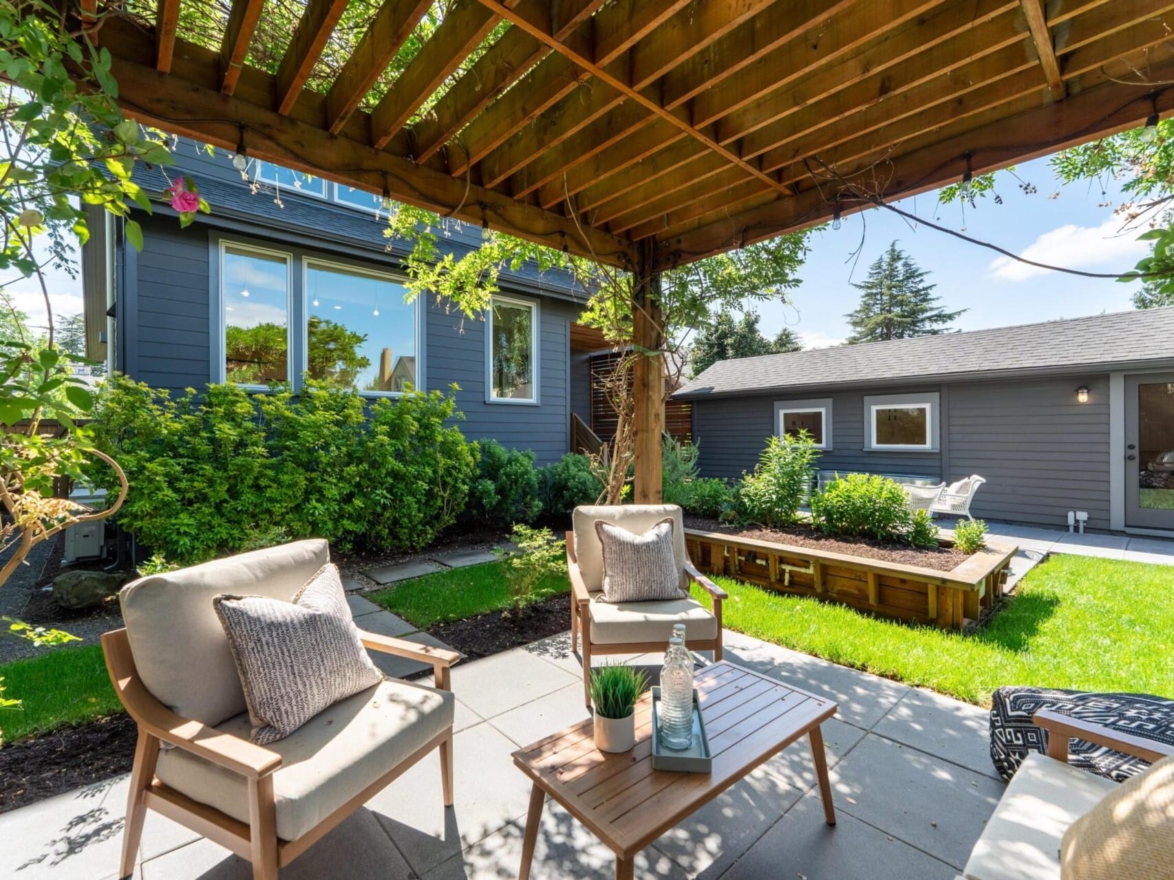 A cozy backyard patio with wooden pergola, cushioned chairs, a small table, and decorative plants. The lush garden includes green shrubs and trees, with a gray house and shed in the background under a bright blue sky.