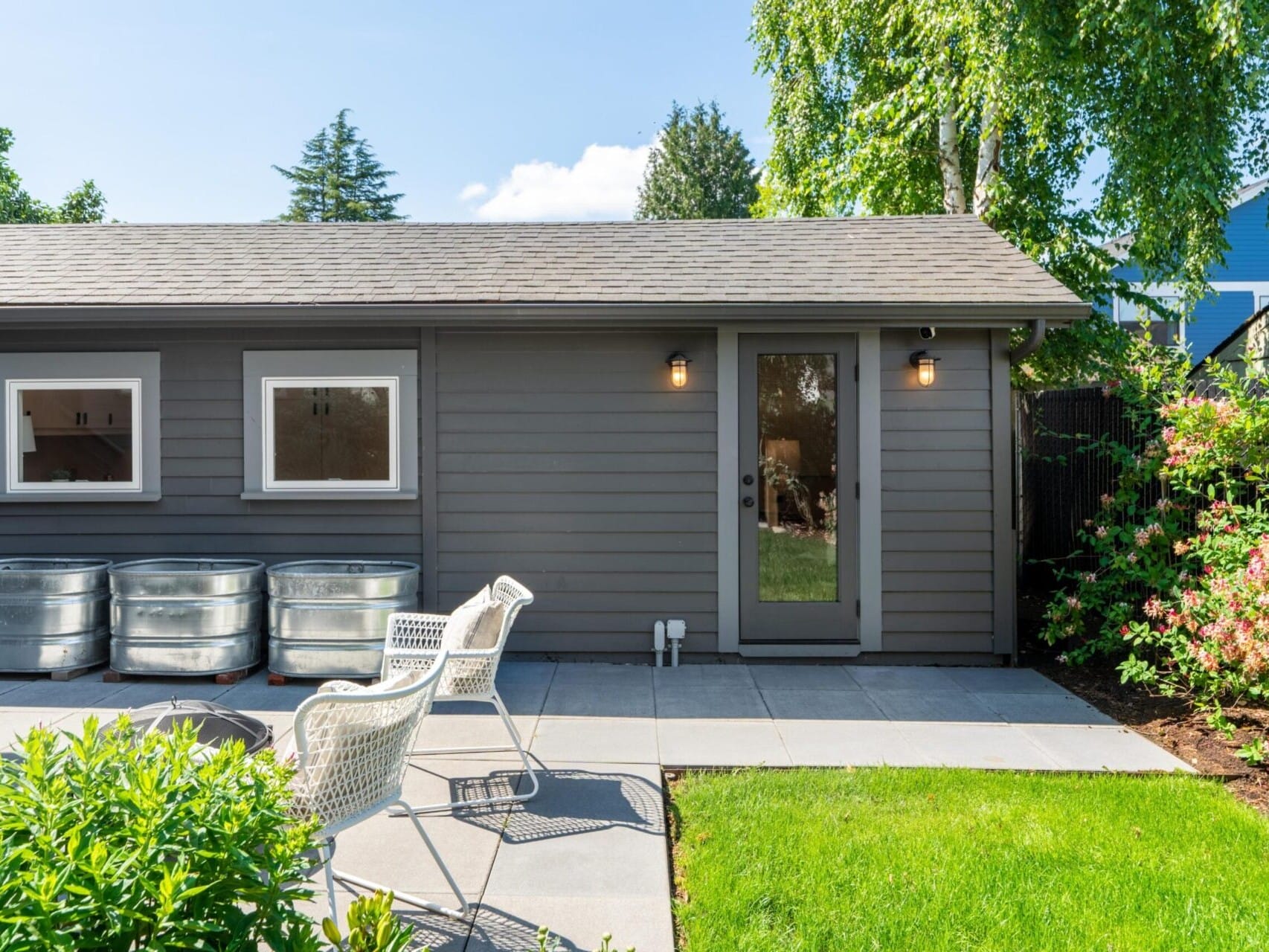 A small gray shed with a glass door and three windows is surrounded by lush greenery. Two white patio chairs are on a concrete slab, next to metal planters. The setting is sunny, with trees and bushes in the background.