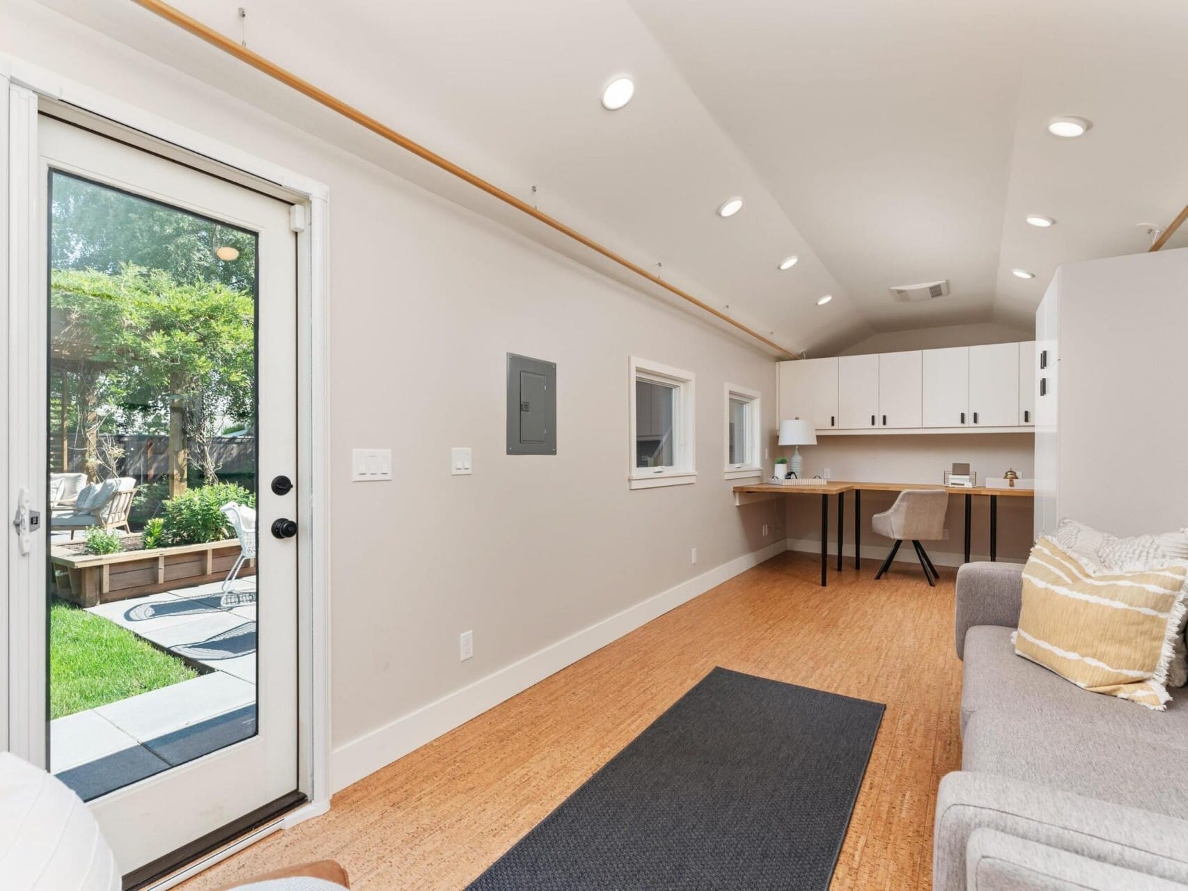 Interior of a bright, modern room with a sofa, desk, and chair. Light wood flooring and a large windowed door lead to a garden. Shelves and cabinets are mounted on one wall, and a rug adds contrast to the minimalist design.