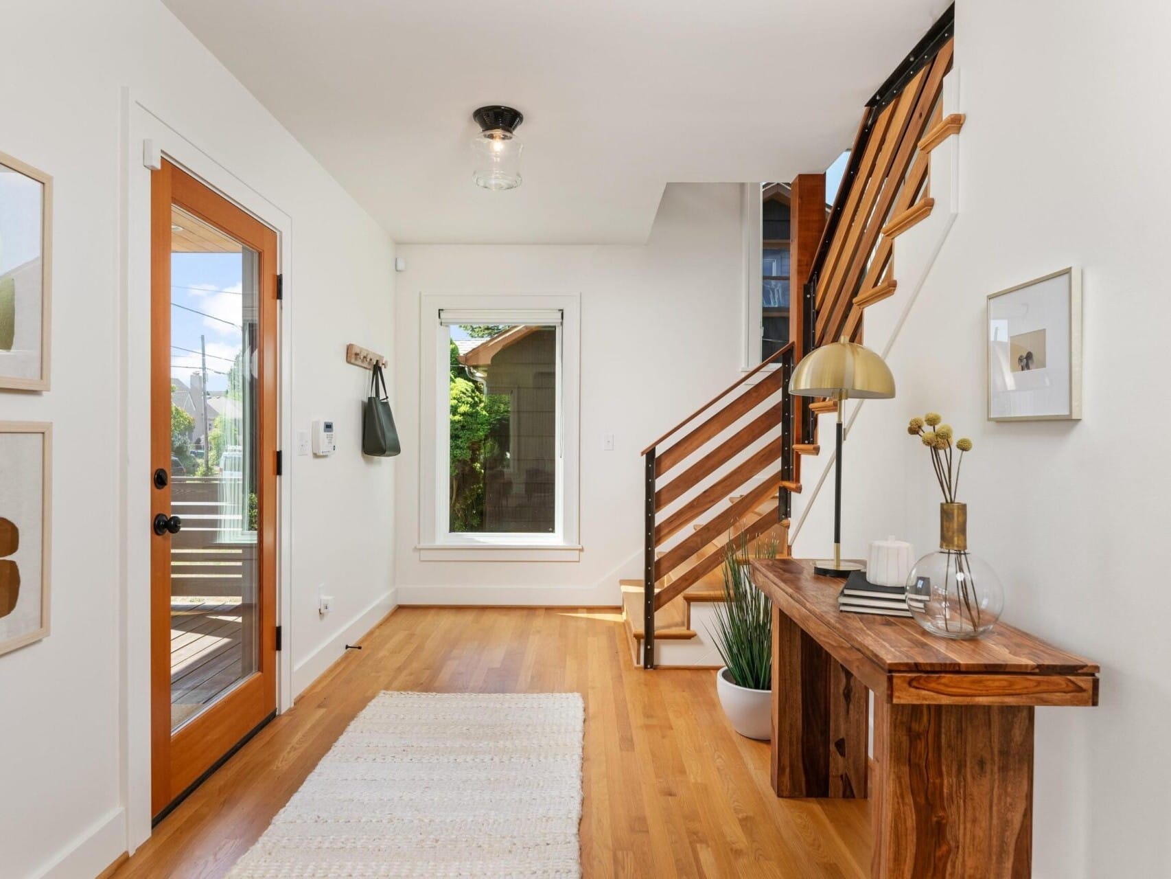 A bright, modern entryway with a wooden floor, a wooden front door with glass panes, and a staircase with wooden railings. A wooden console table holds decorative items. Wall art and a window add to the airy, minimalist decor.