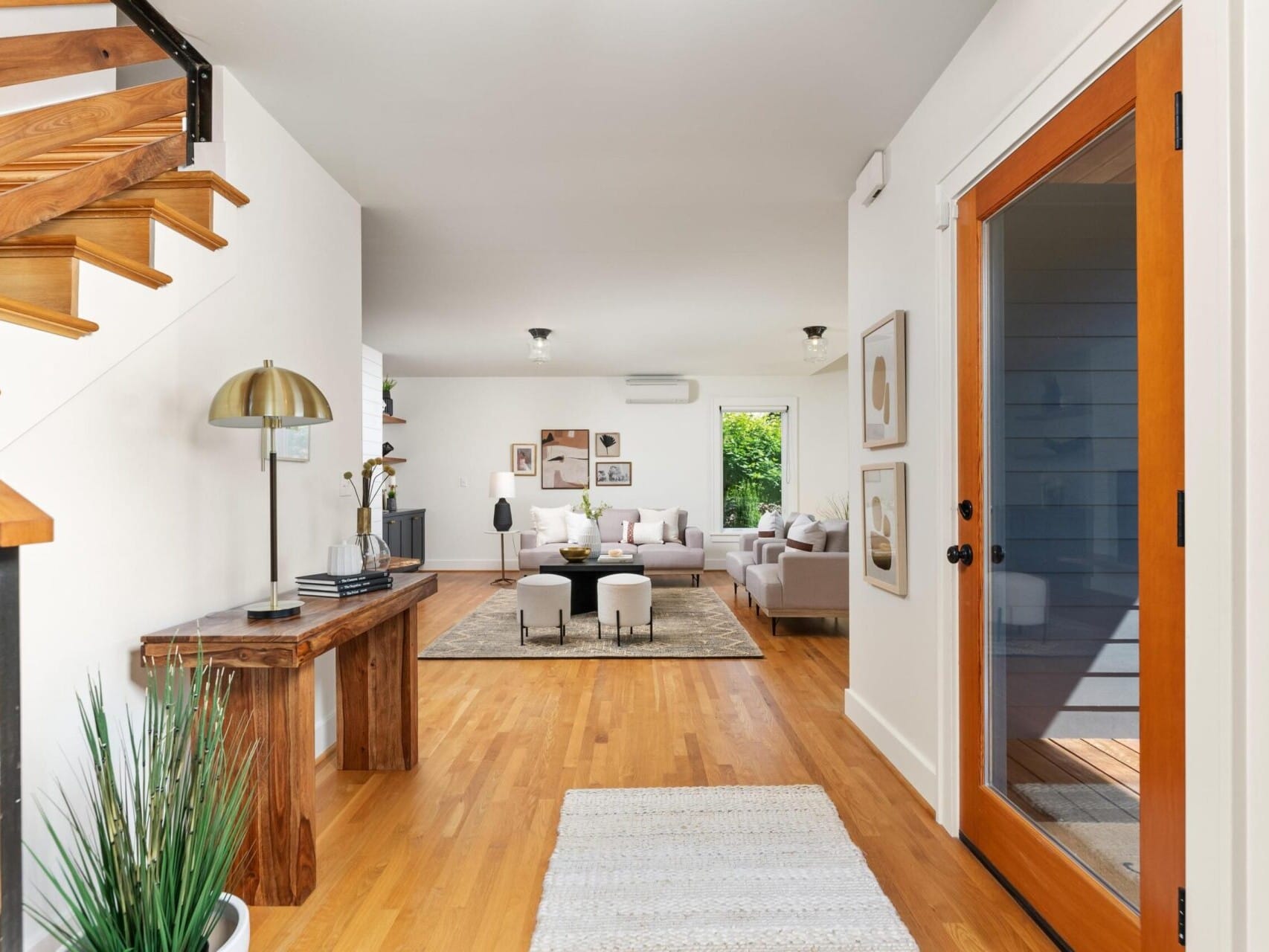 Bright and airy modern living room with wooden floors, a wooden staircase, and a cozy seating area. The entrance features a wooden console table, decorative potted plant, and a large window with a view of greenery outside.