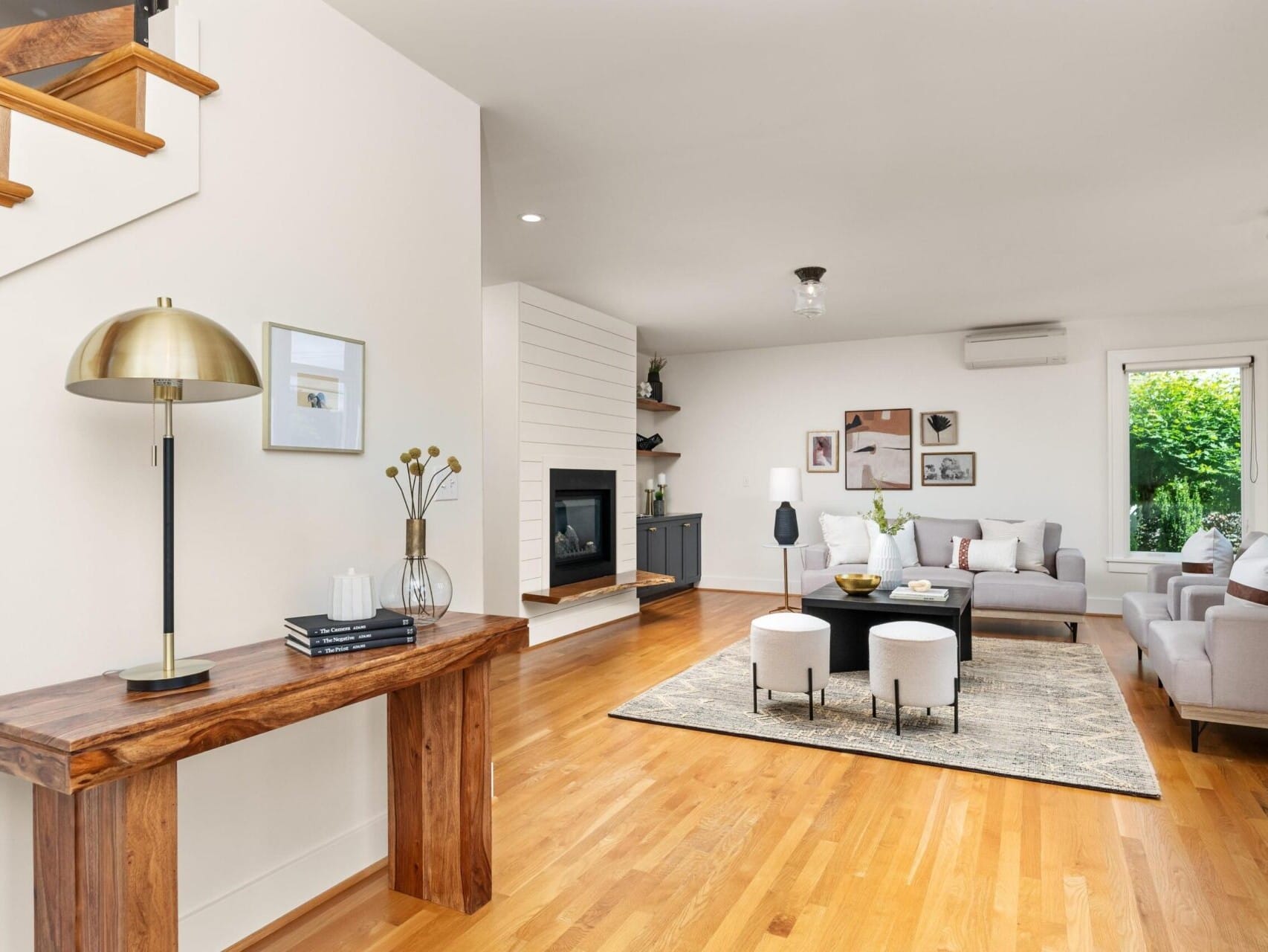 A modern living room with wooden flooring, a light gray sectional sofa, a black coffee table, and a fireplace. The room is decorated with framed art, a brass table lamp, and shelves with plants. A large window provides natural light.