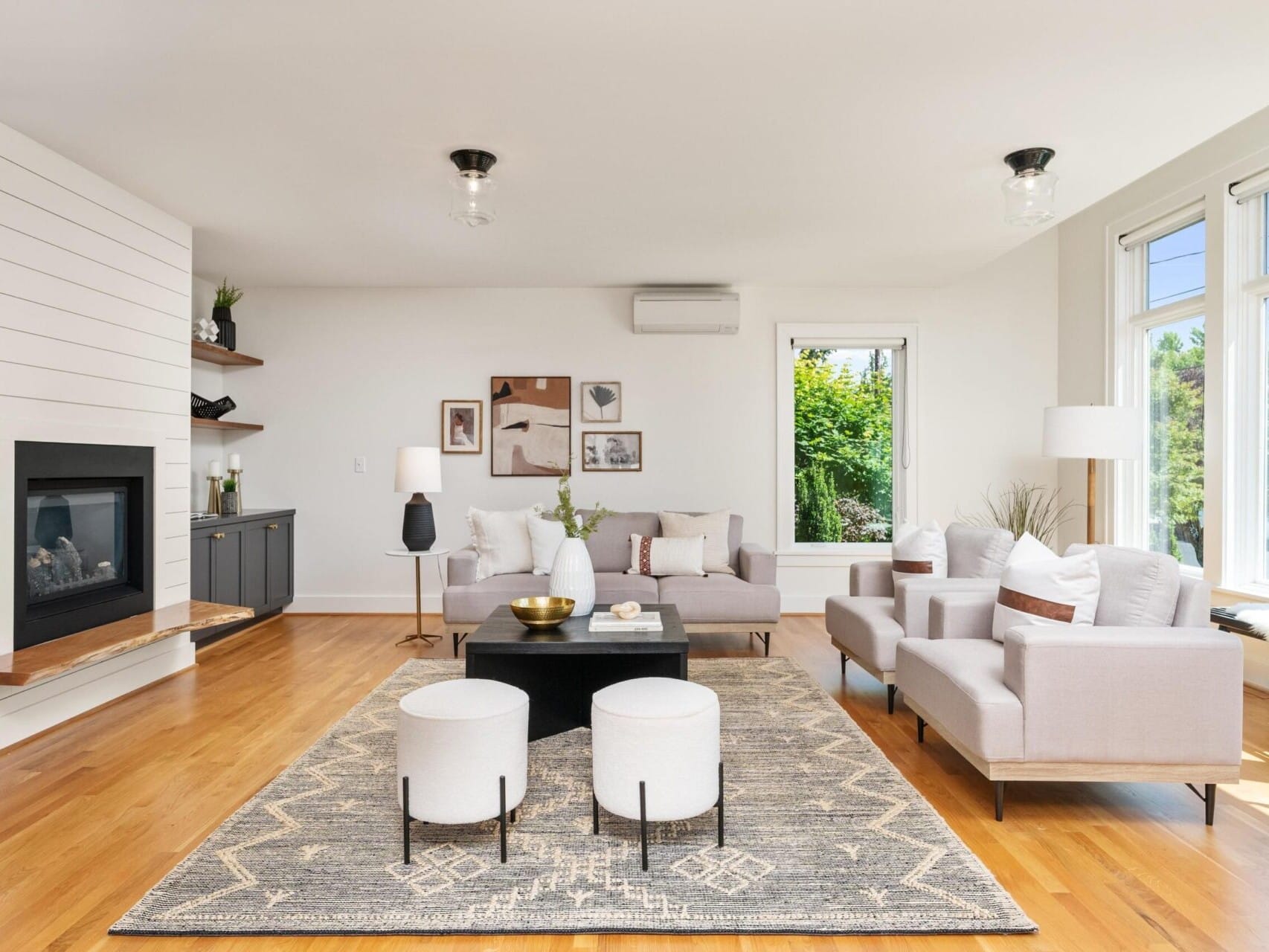 A bright, modern living room features a beige sofa and chairs, a black coffee table with decorative items, two white round stools, and a patterned rug. A fireplace is integrated into a shiplap wall with shelves, and large windows offer an outdoor view.