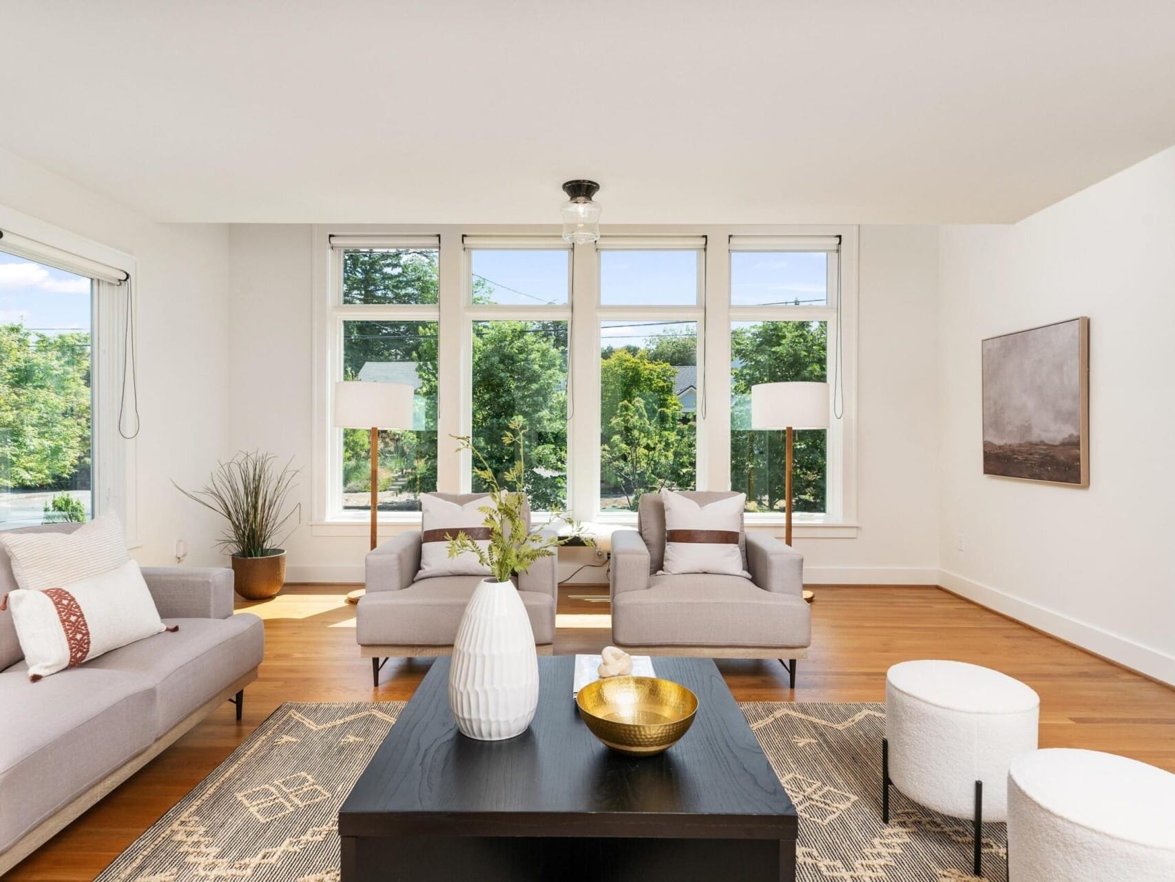 A bright, modern living room with large windows, two beige armchairs, a gray sofa, and a black coffee table. Decor includes a white vase with flowers and a golden bowl. Two white poufs and floor lamps are also visible, with greenery outside.