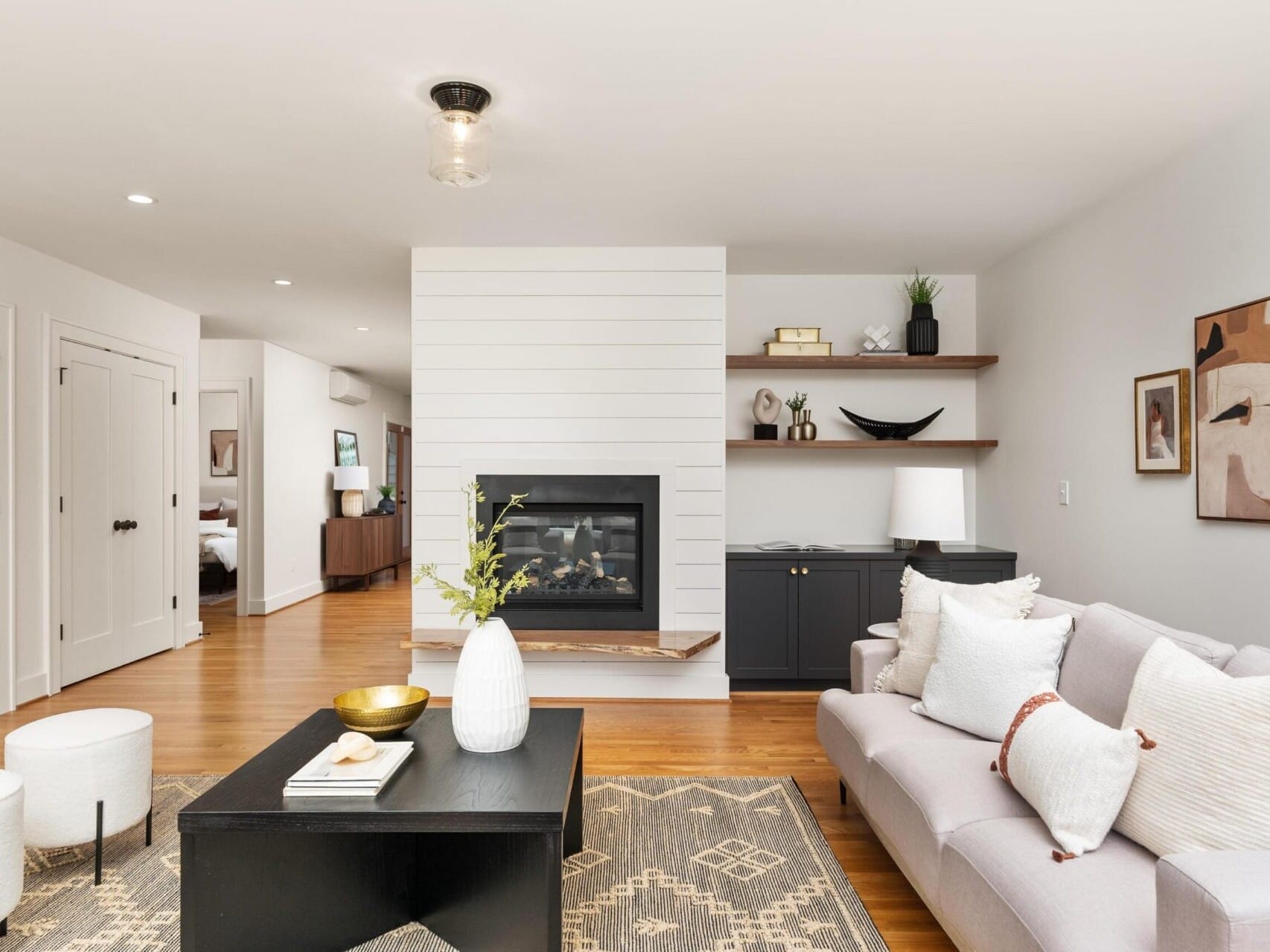 A modern, cozy living room with a light beige sofa, a patterned rug, and a black coffee table. A fireplace is set into a white paneled divider, with shelves and cabinets on one side. The space features neutral colors and minimalistic decor.