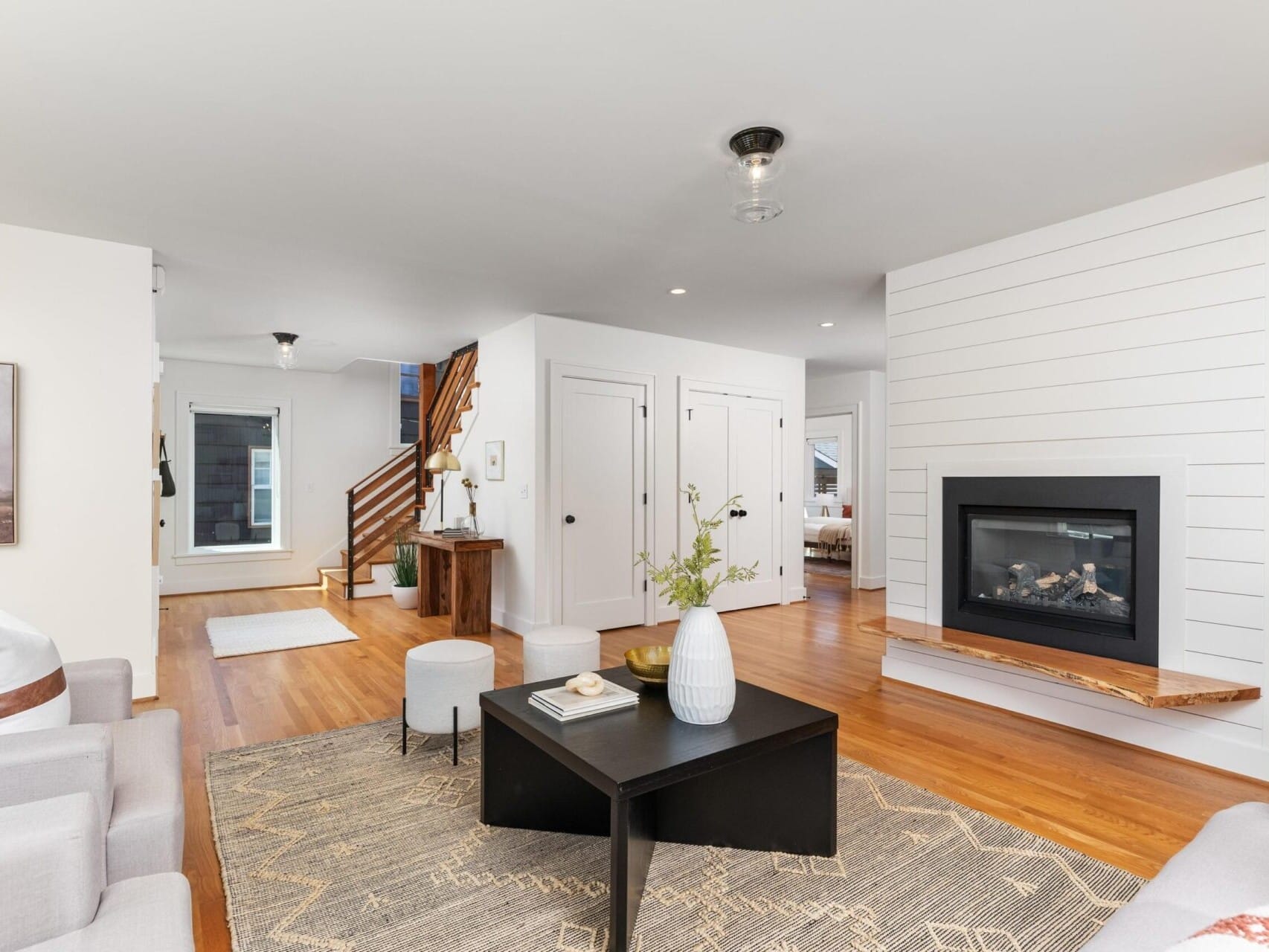 Modern living room with hardwood floors, a black coffee table, and light gray sofas. A gas fireplace with shiplap surround is on the right. A staircase with wooden steps and a small console table is in the background. Neutral décor and soft lighting.