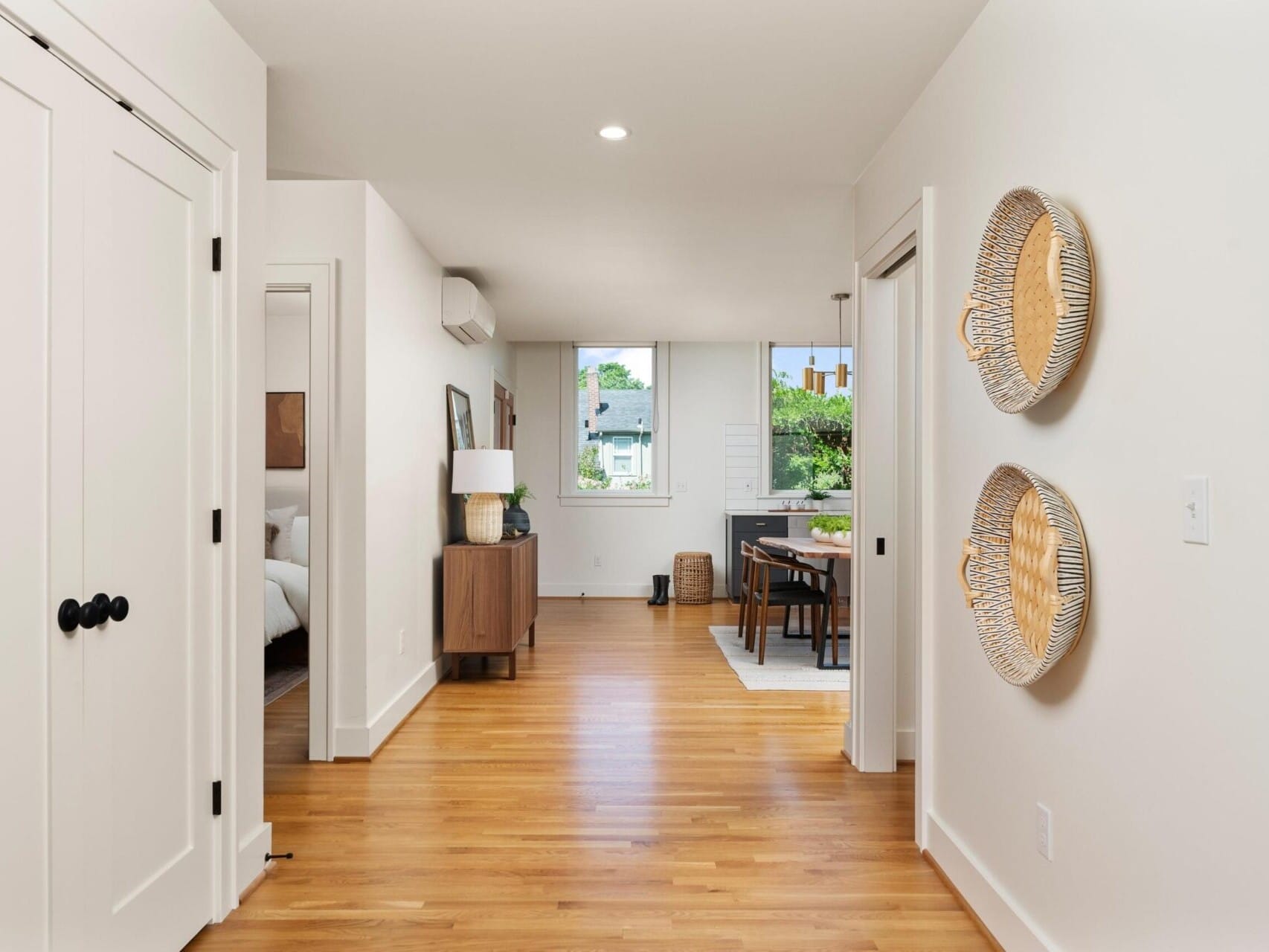 A bright, modern hallway with wooden flooring leads to an open-plan living area. A bedroom is visible on the left, decorated with neutral tones. Two decorative plates hang on the wall, and a small round dining table is placed near a large window.