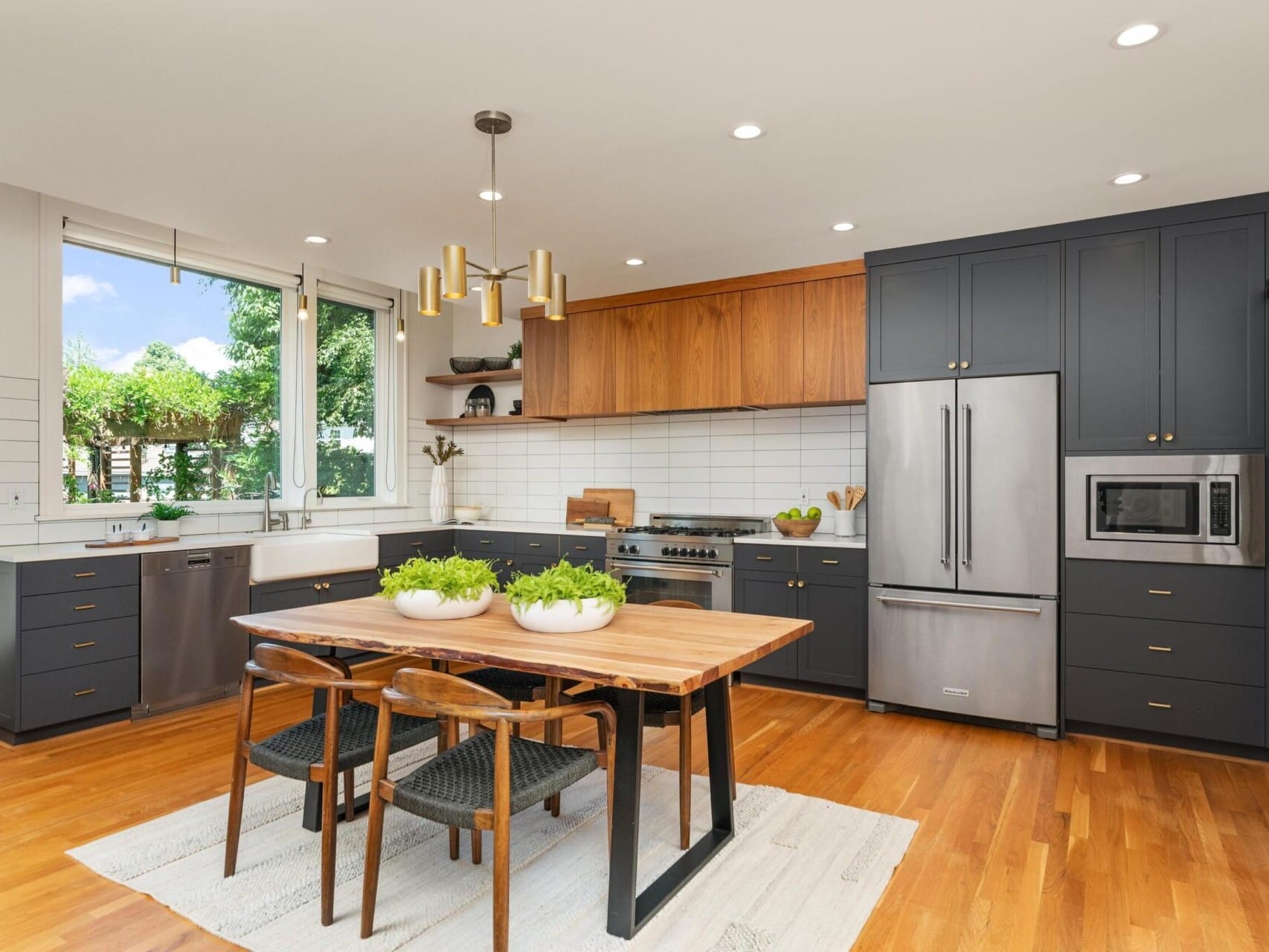 A modern kitchen with dark cabinets, a wooden dining table, and black chairs. Stainless steel appliances, including a refrigerator and oven, are integrated. Large window provides natural light. Potted plants are on the table. Hardwood floors throughout.