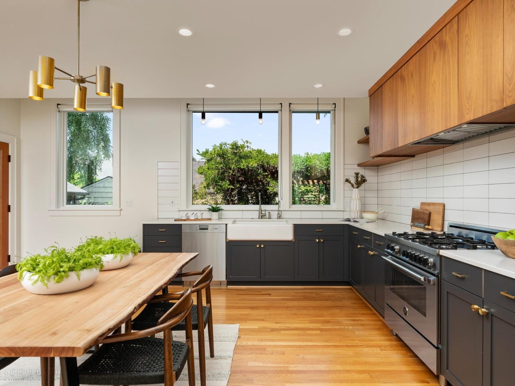 A modern kitchen with wood flooring, white walls, and a central wooden dining table with potted plants. Black lower cabinets and wooden upper cabinets line the walls, and a window offers a view of greenery outside. Stylish lighting hangs above.