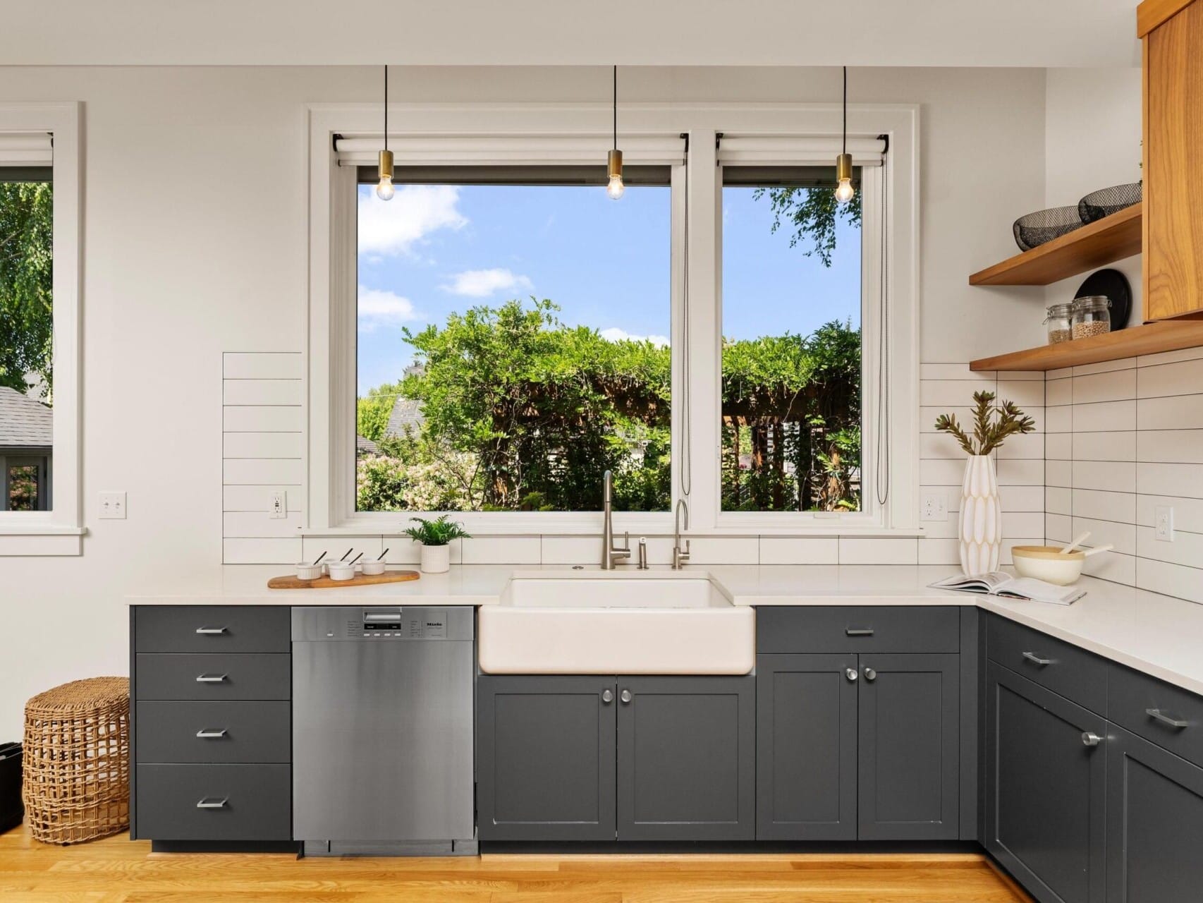 Modern kitchen with dark gray cabinets and a farmhouse sink under a large window. White subway tiles line the walls. Natural light illuminates wooden shelves and a stainless steel dishwasher. A woven basket and boots are in the corner.