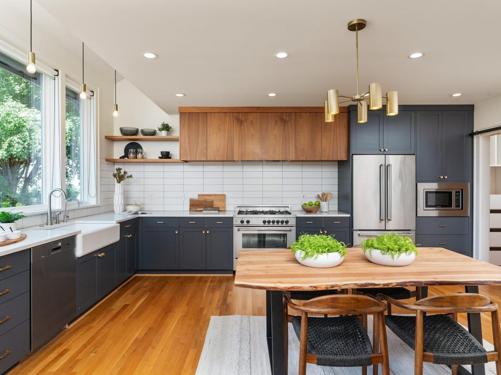 A modern kitchen with a wooden dining table and chairs in the foreground. The room features blue cabinets, a white backsplash, stainless steel appliances, and a farmhouse sink. Plants are placed on the table and shelves, and large windows let in natural light.